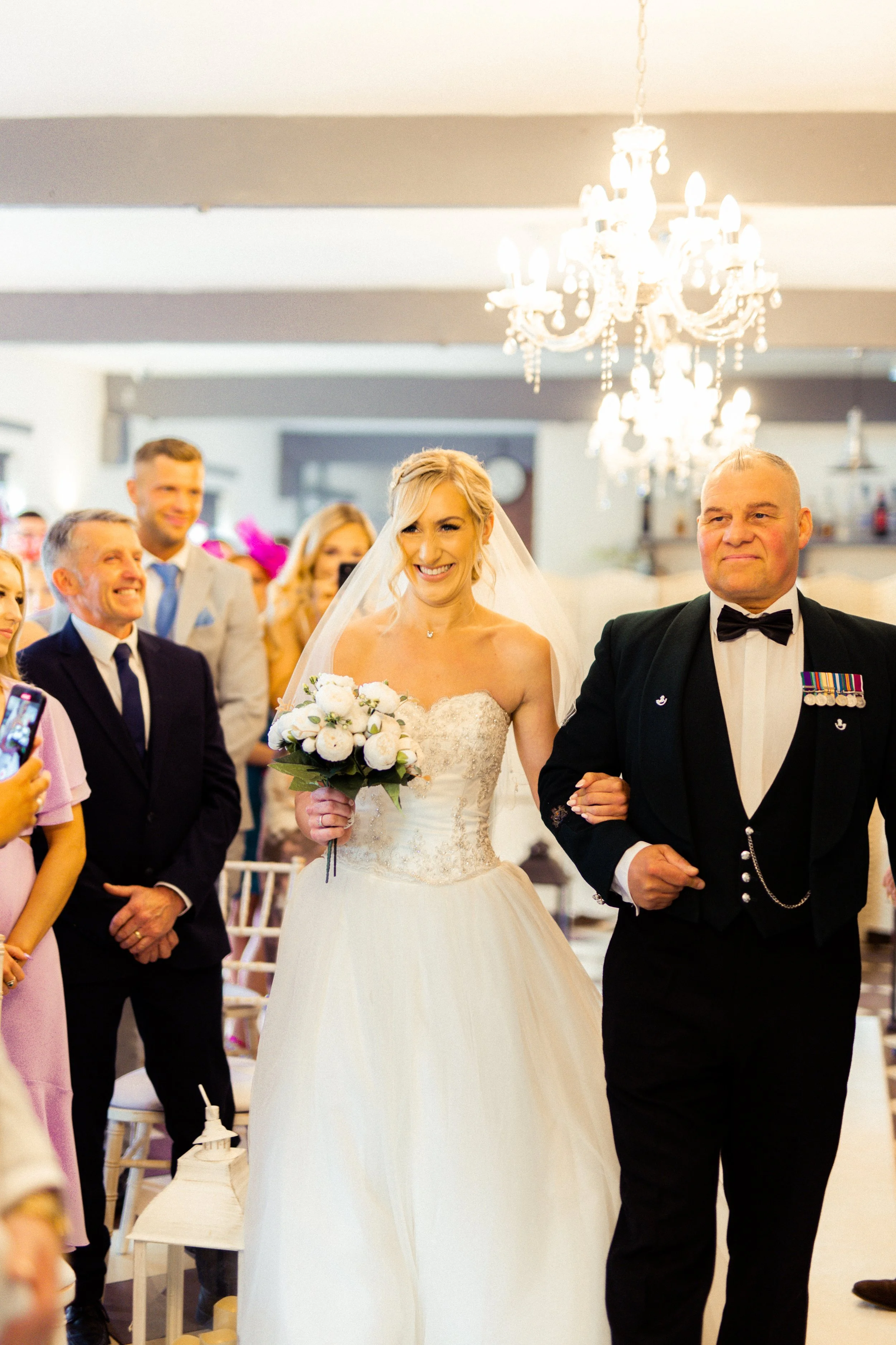 A bride walking down the aisle with her escort at a wedding ceremony, surrounded by smiling guests in an indoor setting with a chandelier overhead.