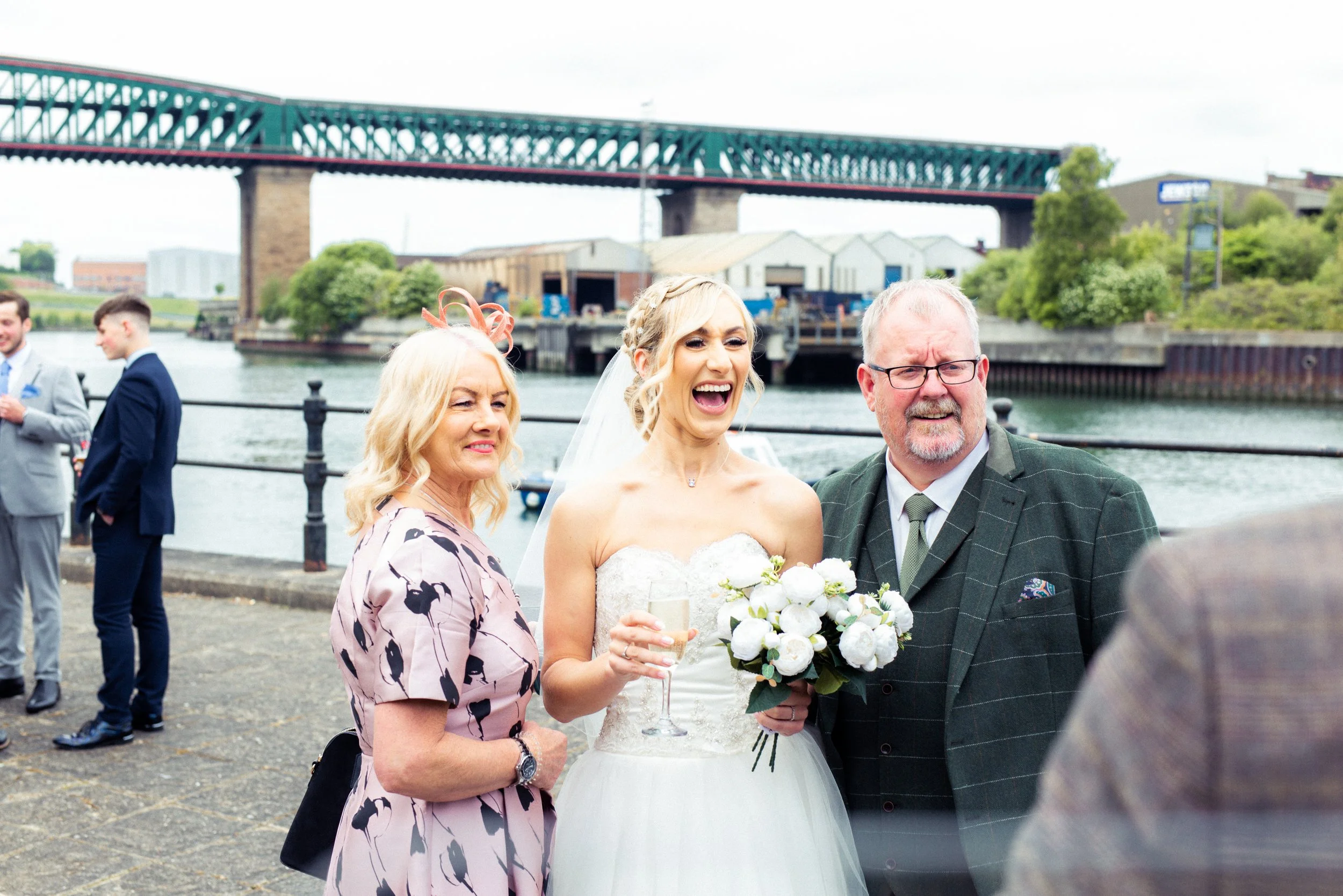 Wedding celebration by a river with a bridge in the background, featuring a bride in a white gown holding a bouquet and champagne, flanked by two guests, one woman in a pink dress and a man in a checkered jacket.
