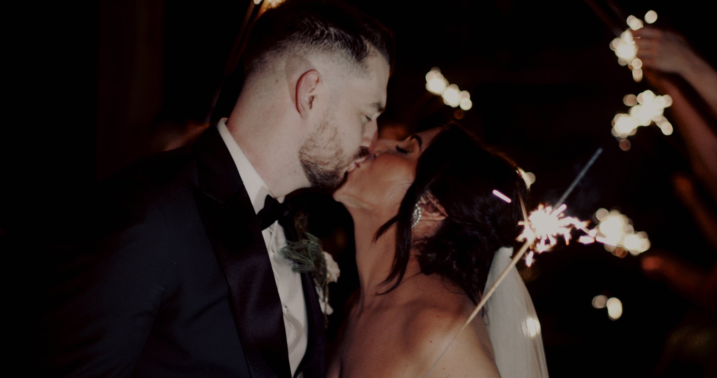 A bride and groom kiss at their wedding reception, with sparklers in the foreground and a dark background.