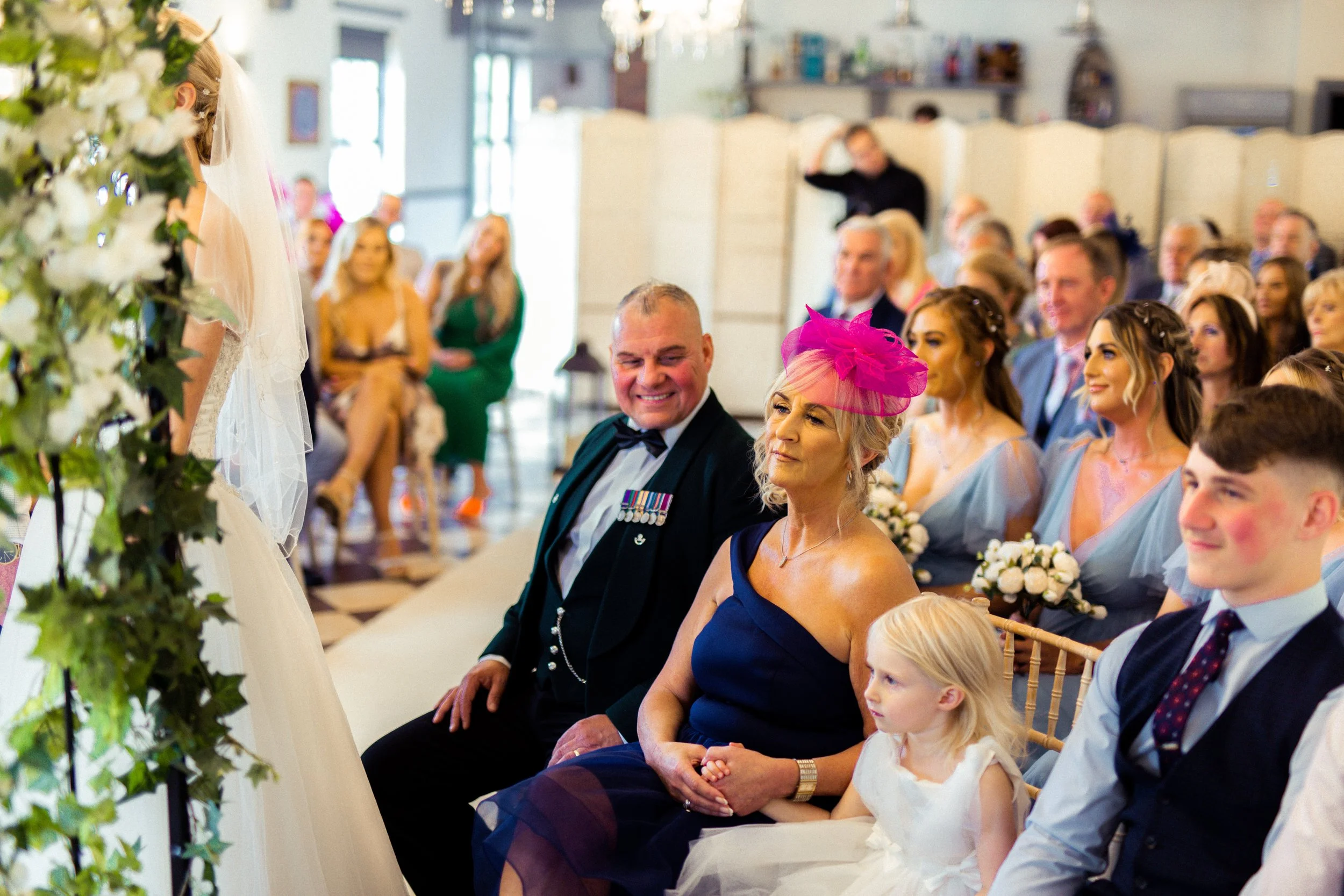 A wedding ceremony with guests seated and a bride partially visible on the left, with white flowers and greenery in the foreground.