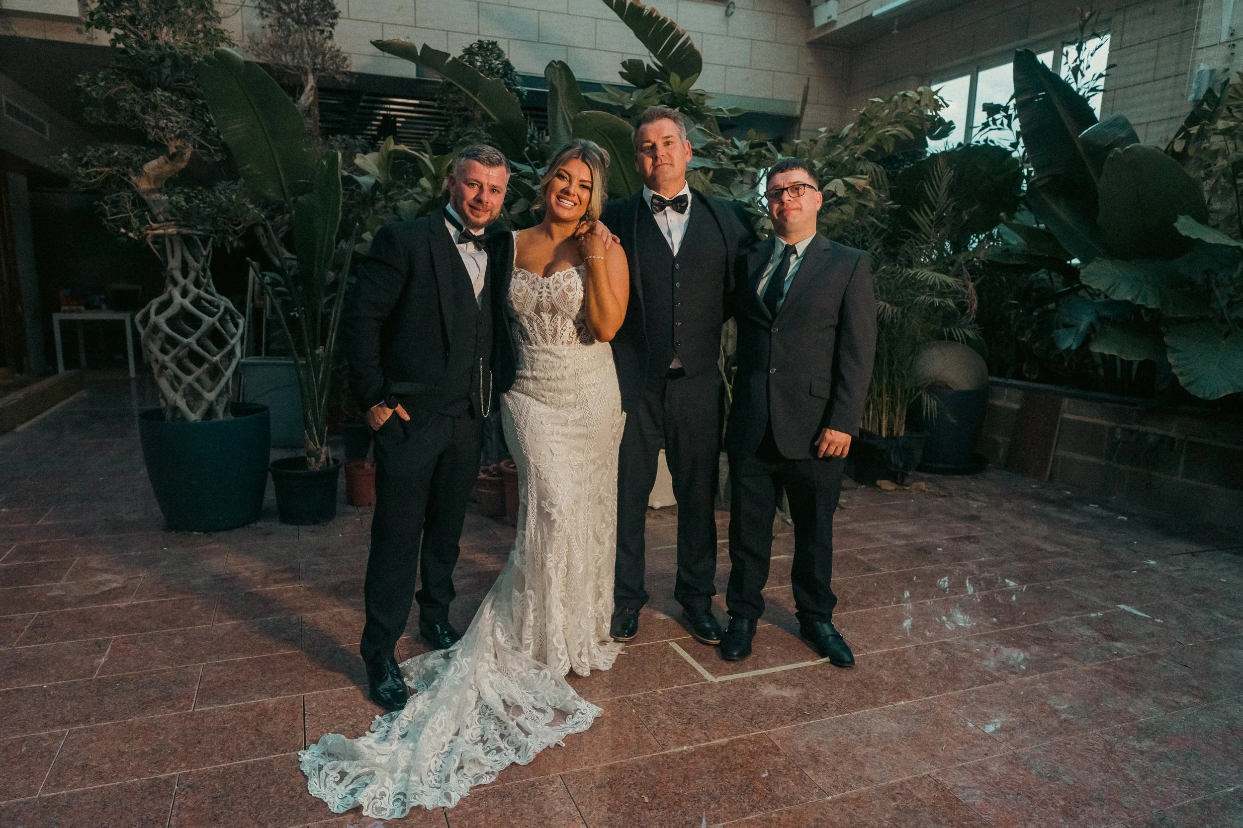 Group of five people dressed in formal attire standing together indoors in front of large plants.