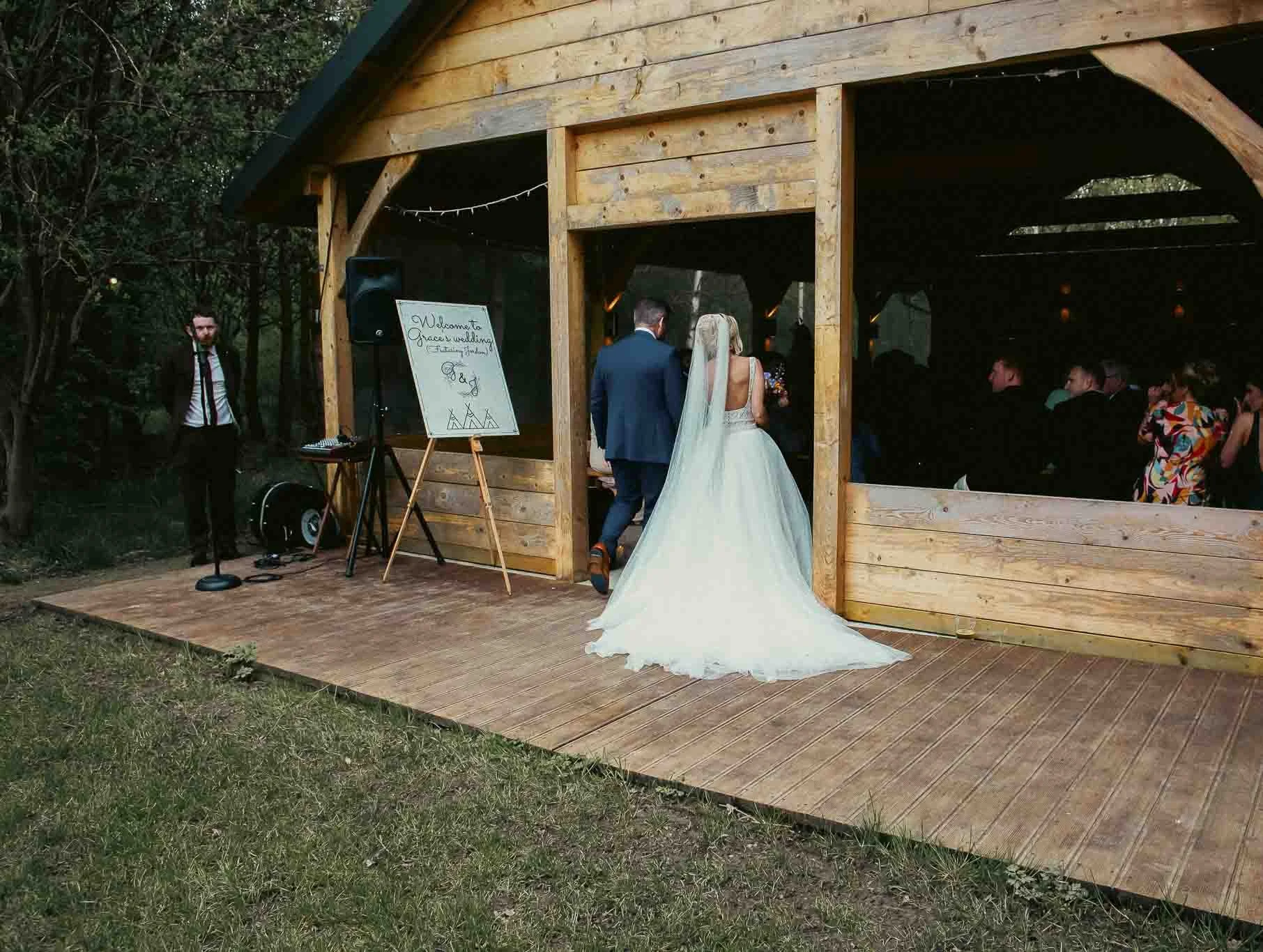 Bride and groom walking into a wedding reception at a wooden outdoor venue with guests seated inside.