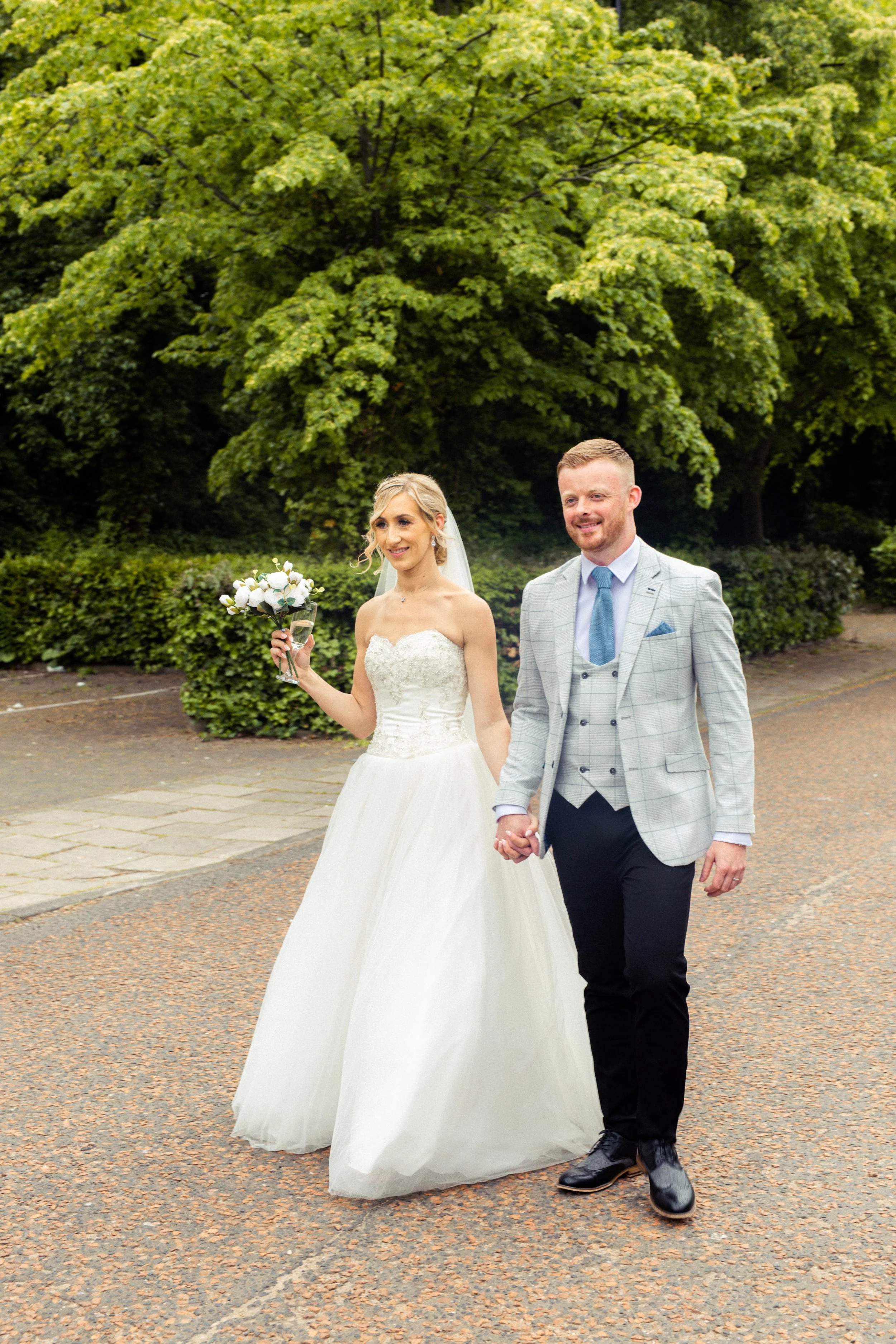 Bride and groom walking hand in hand outdoors, woman in white wedding gown holding a bouquet, man in gray plaid suit, trees in background.