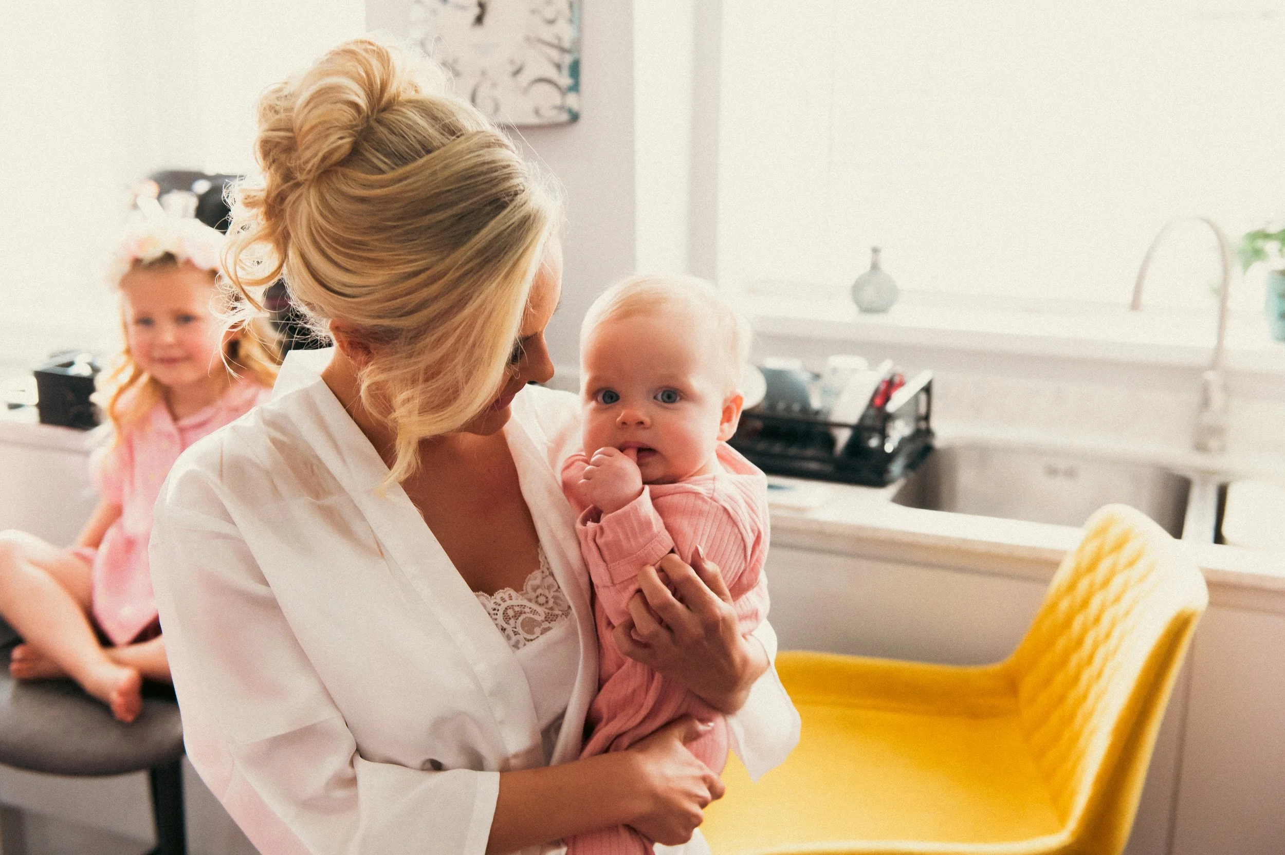 A woman holding a baby girl with a hand in her mouth while standing in a bright, modern kitchen, with another girl sitting on a chair in the background.