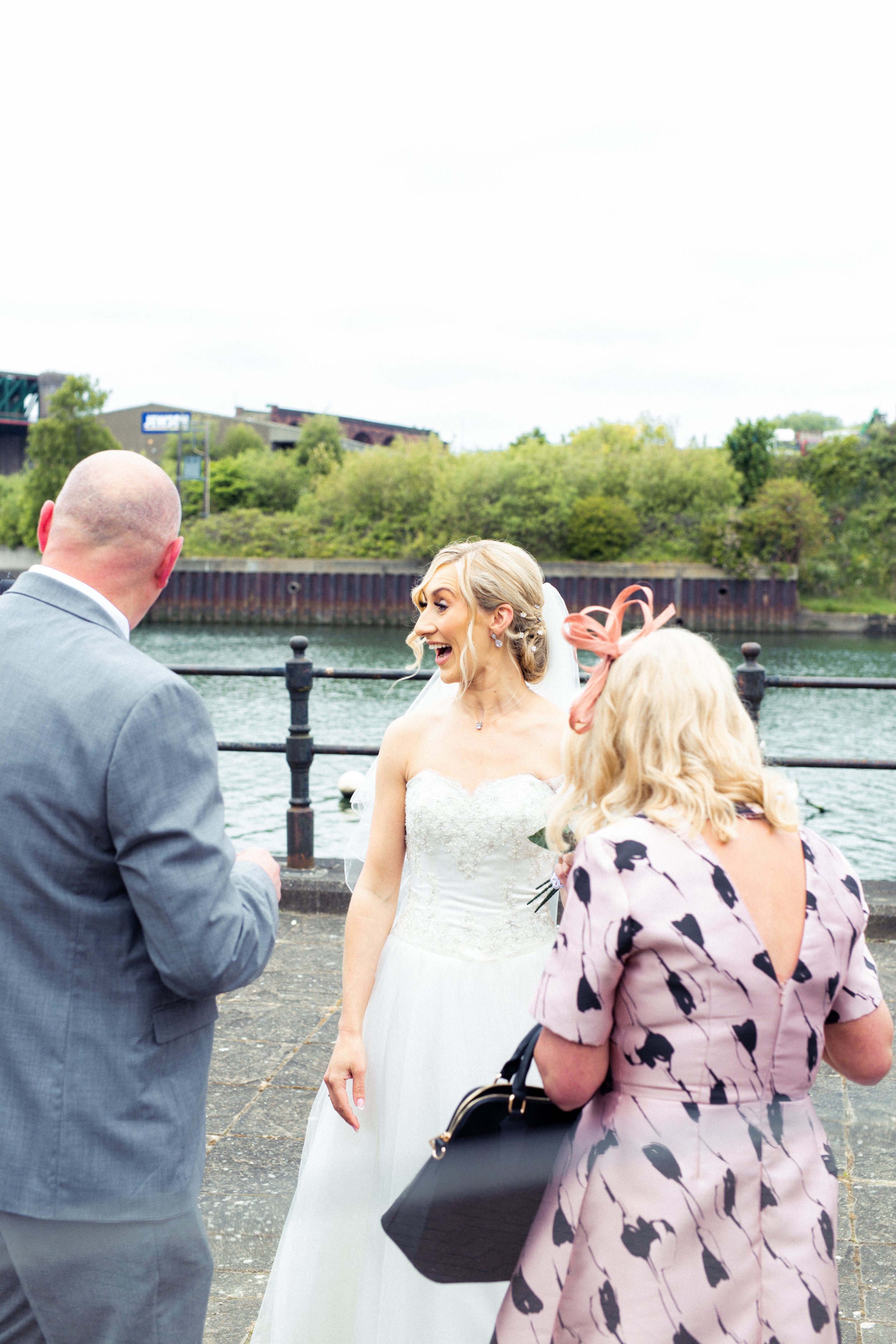 A bride with blonde hair and a strapless lace wedding gown smiling and talking to a man in a gray suit and a woman with blonde hair wearing a pink dress with black animal prints and a large headband, near a river with a cityscape and greenery in the 