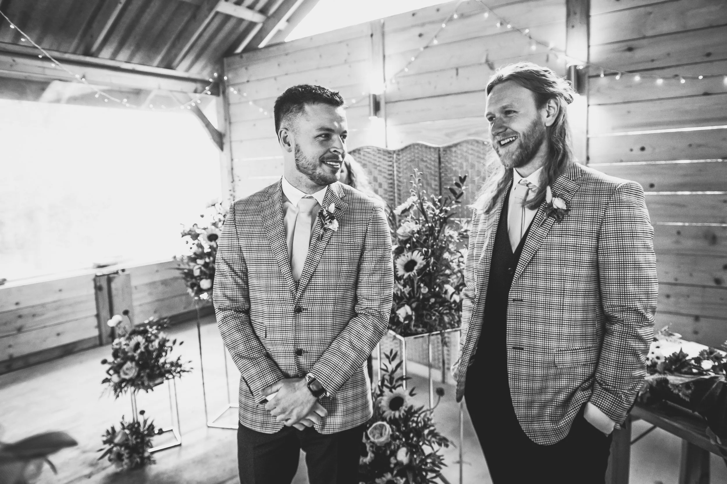 Two men in checkered suits, smiling and talking, standing indoors with wooden walls, floral arrangements, and string lights.