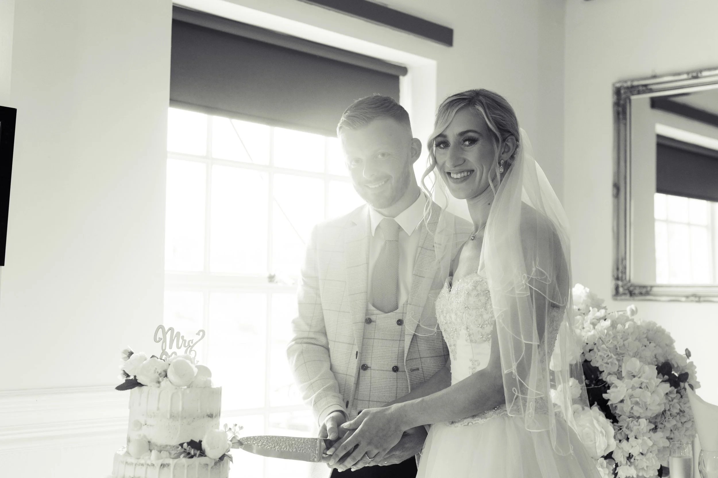 A bride and groom smiling while cutting a wedding cake at their wedding reception inside a bright room with large windows and a mirror on the wall.