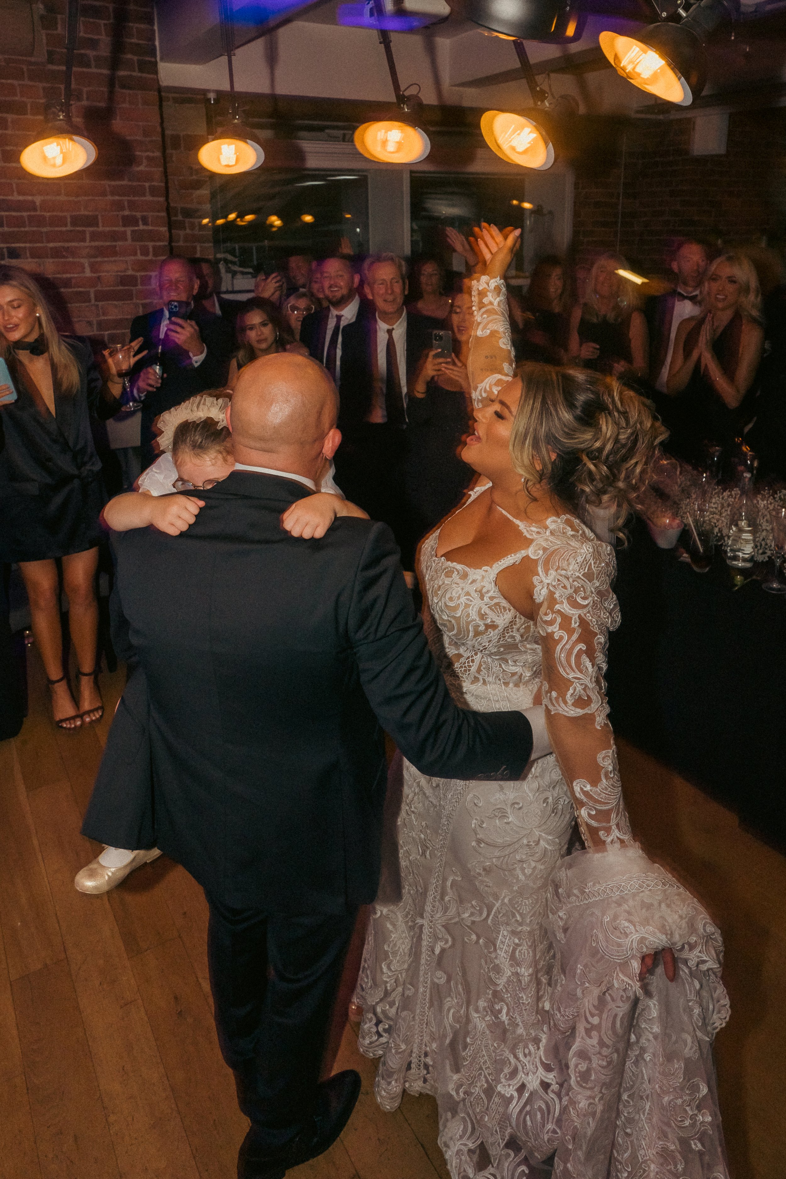 A wedding reception with the bride and groom dancing, surrounded by guests. The bride is wearing a lace wedding dress and the groom is in a black suit. Guests are smiling, clapping, and taking photos in a warmly lit venue with brick walls.