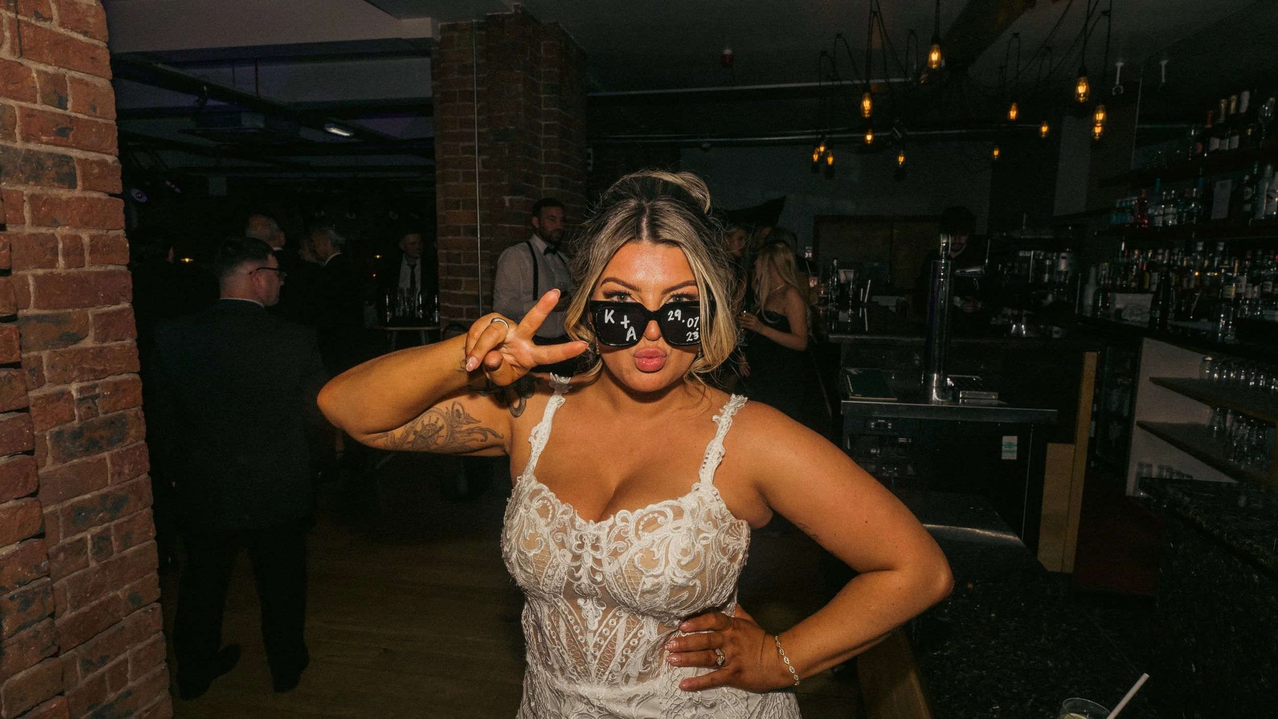 A woman in a white lace dress wears black sunglasses with writing on them, making a peace sign and posing with her lips puckered inside a dimly lit bar.