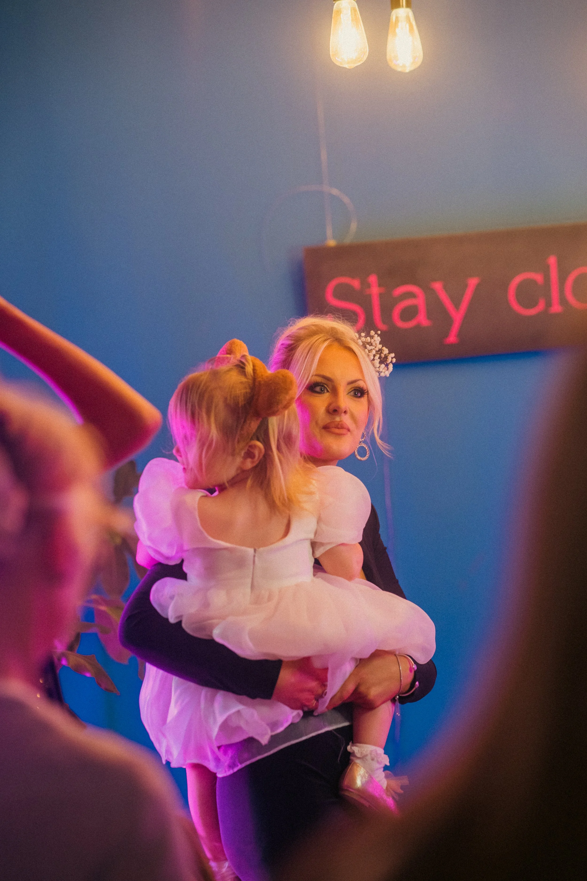 A woman holding a young girl in a ballet costume at an indoor event. The woman has blonde hair and is wearing earrings, and the girl has blonde hair with bear ears headband, wearing a pink tutu dress. There is a sign in the background that reads "Sta