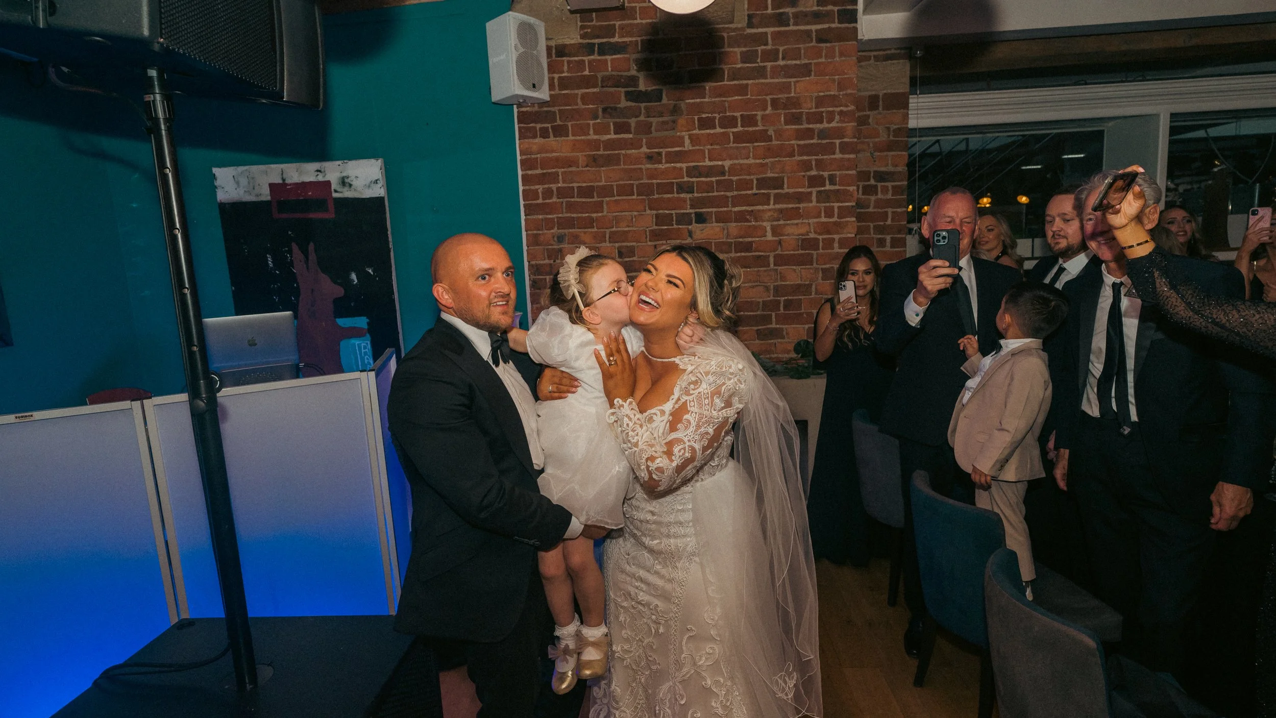 A bride in a lace wedding dress and veil is happily hugging a young girl, who is kissing her cheek. The groom is holding the girl, dressed in a suit with a bow tie. In the background, several guests in formal attire are taking photos and watching the