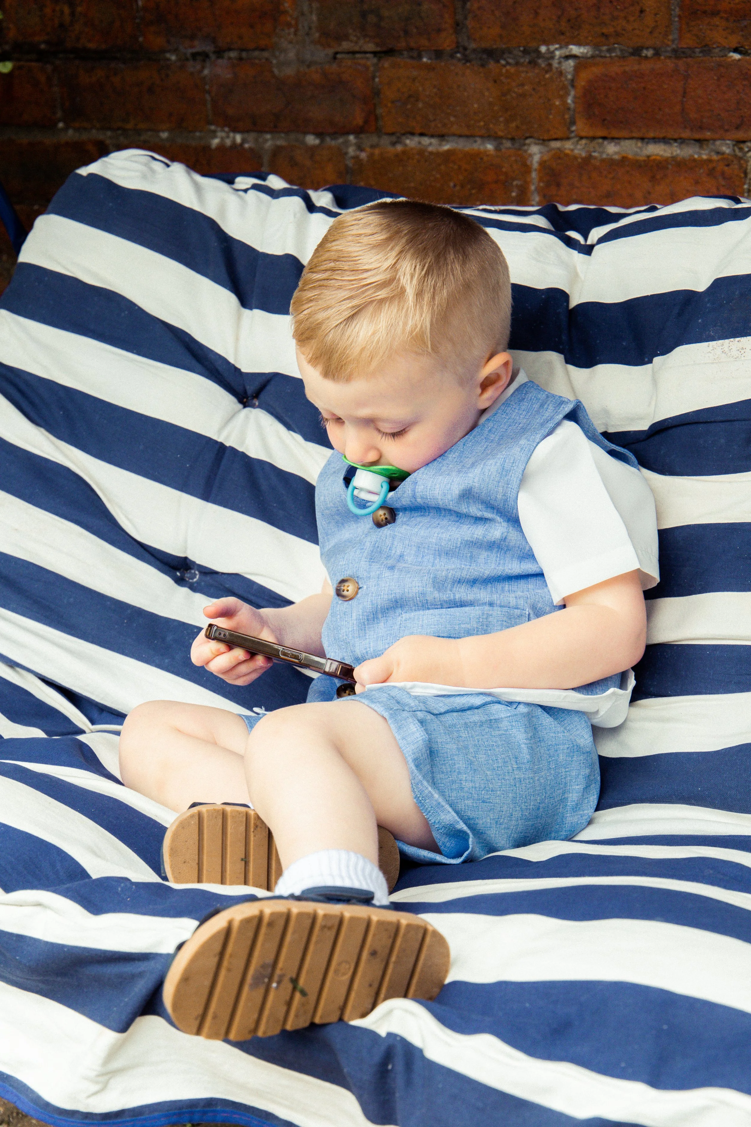 A young boy sitting on a striped blue and white couch, looking at a smartphone with a pacifier in his mouth.