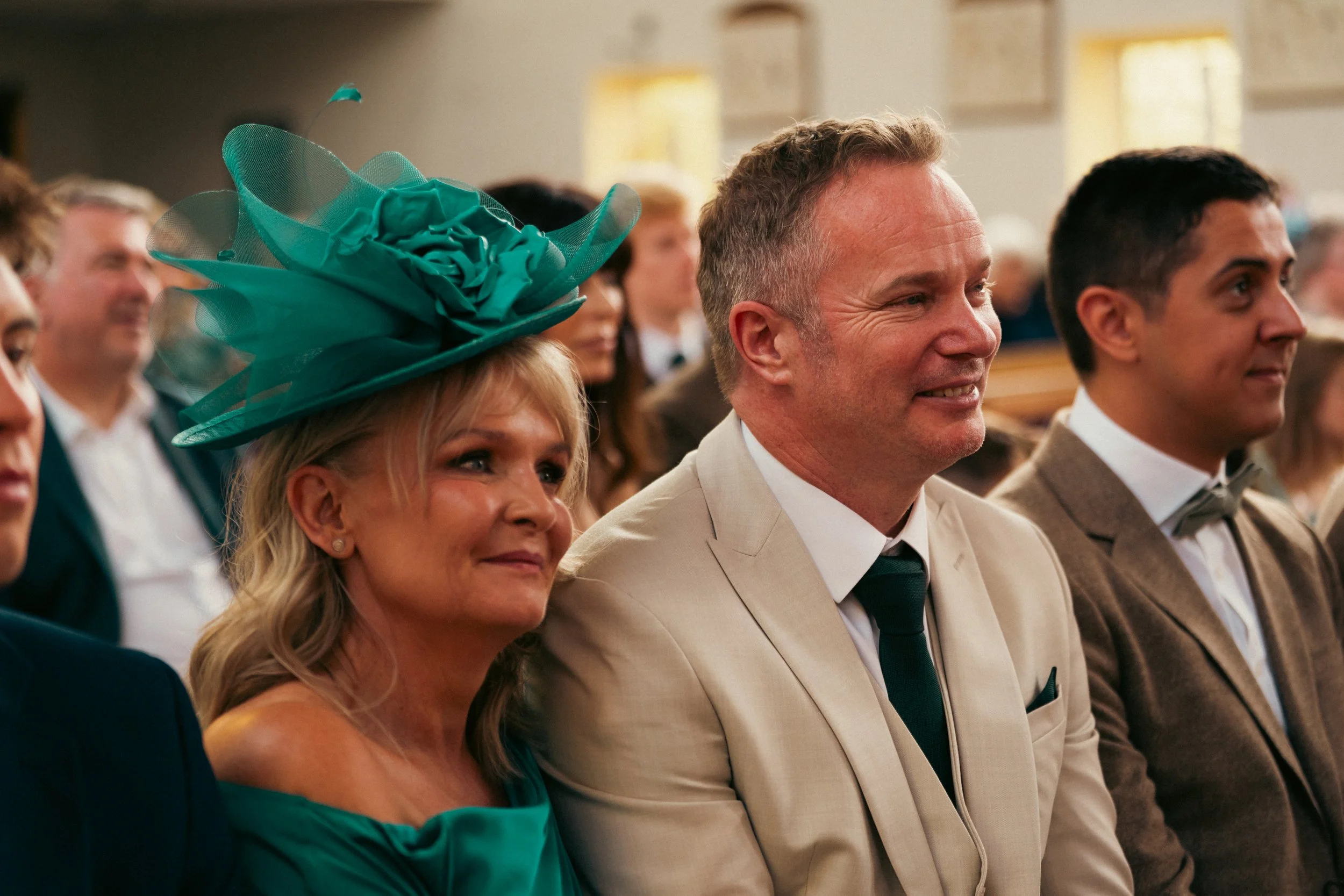 People attending a formal event, with a woman wearing a large teal hat and teal dress sitting next to a man in a cream suit and dark green tie, in a church or similar setting.