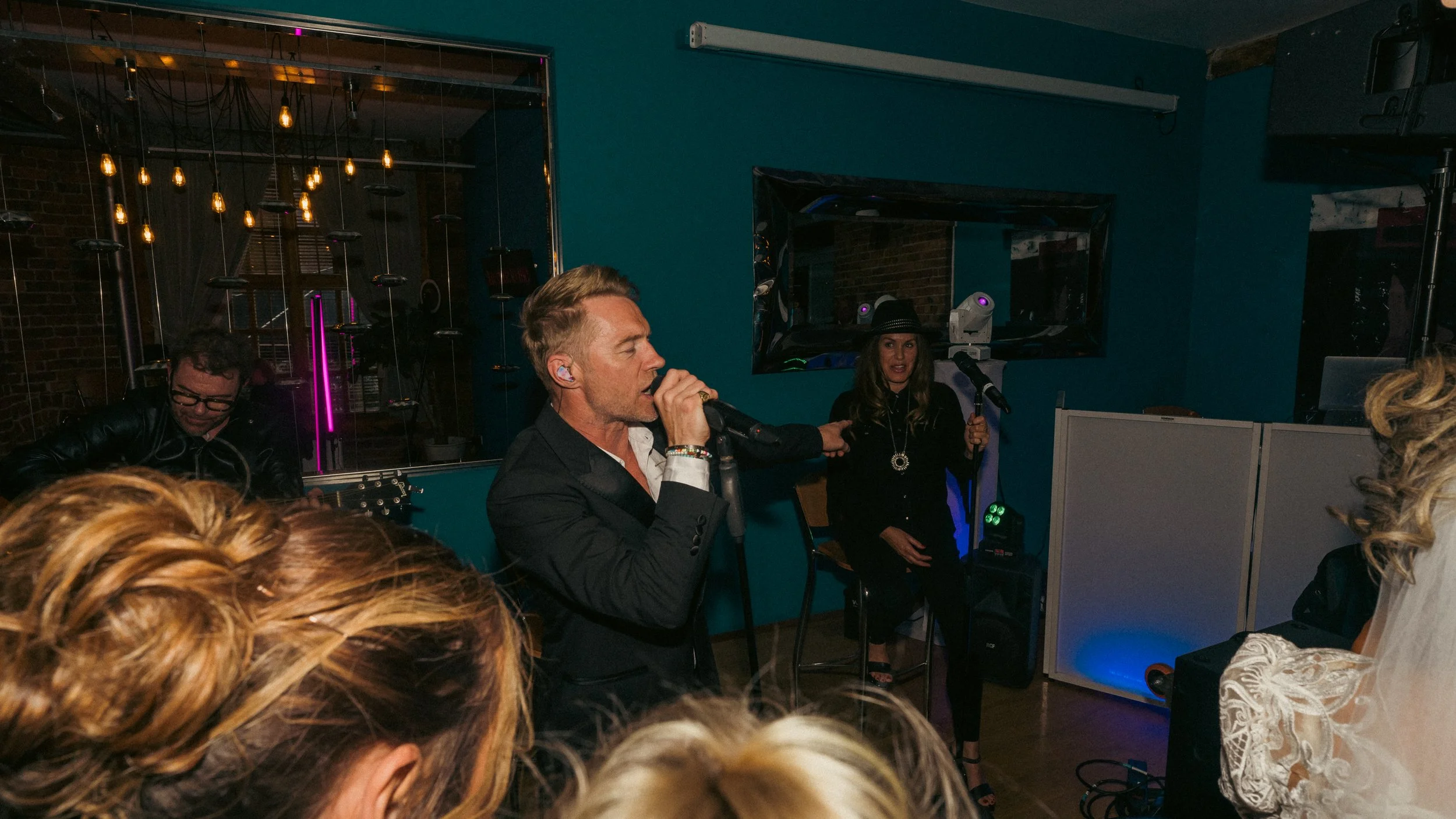 A group of people performing at a karaoke night in a cozy, dimly lit bar with blue walls, with a male singer wearing a black suit and two women, one with a black hat, singing along with microphones, while others watch and listen.