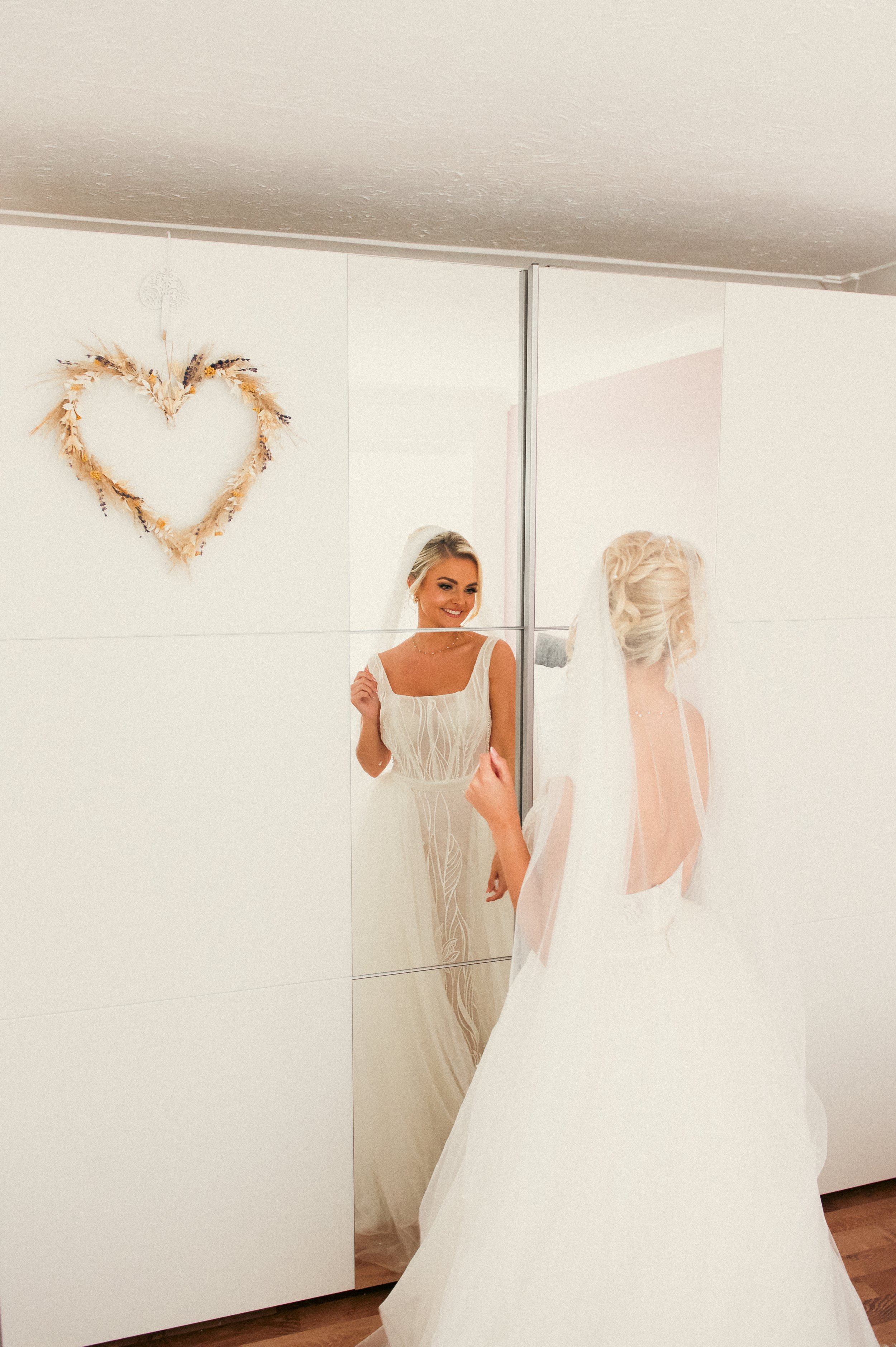 Bridal woman in white dress with veil looking at herself in a mirror, heart-shaped decoration made of dried flowers on the wall.