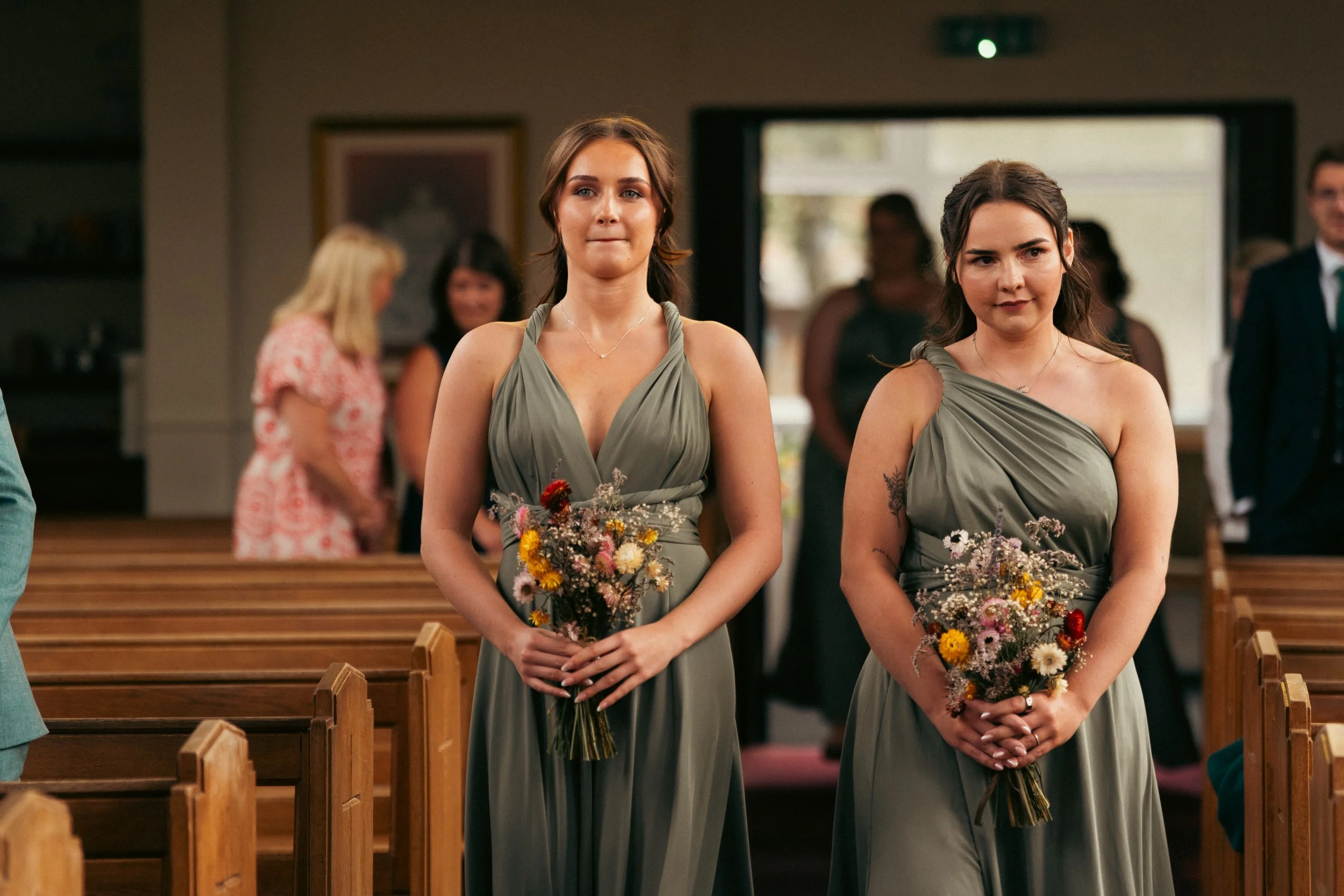 Two women wearing matching gray bridesmaid dresses standing inside a church, holding small bouquets of mixed flowers, during a wedding ceremony.