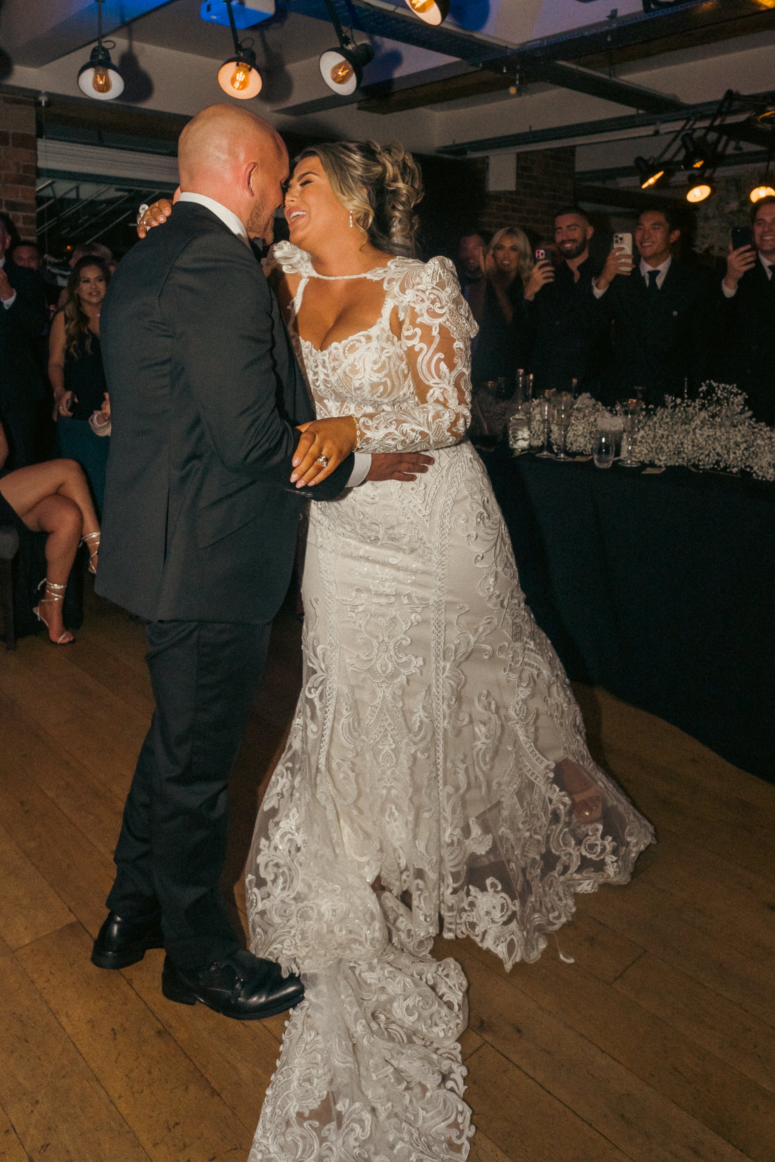 A bride and groom dancing at their wedding reception, surrounded by guests who are taking photos and watching happily.