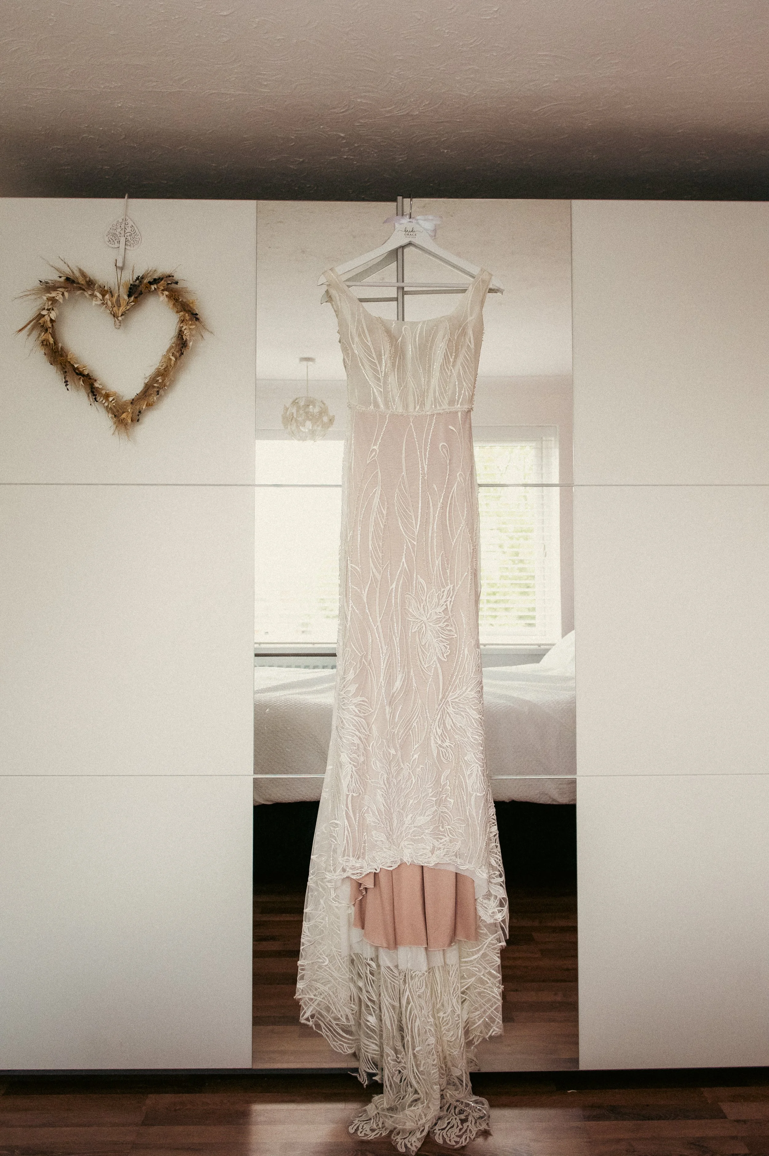 A wedding dress hanging on a hanger in front of a mirror, with a heart-shaped wreath on the wall to the left.