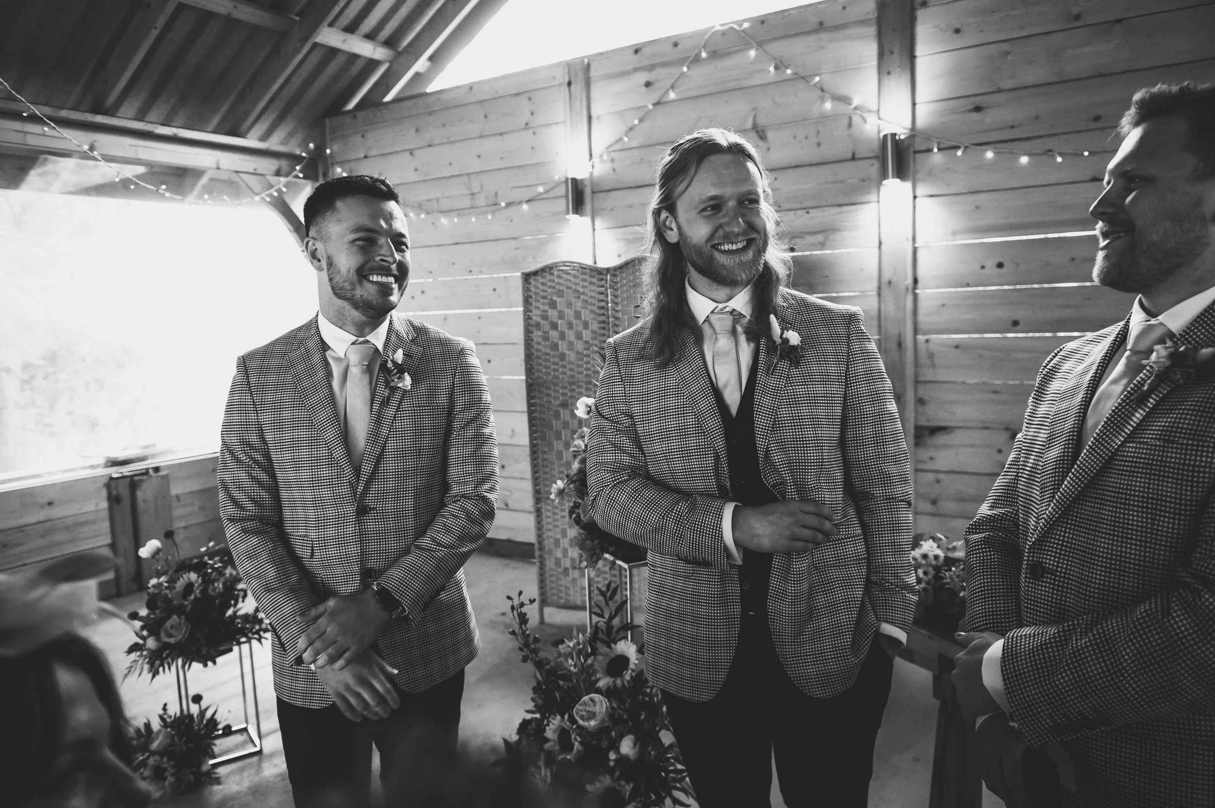 Black and white photo of four men in tuxedos with boutonnieres, smiling and talking in a rustic wooden room decorated with string lights and flowers.