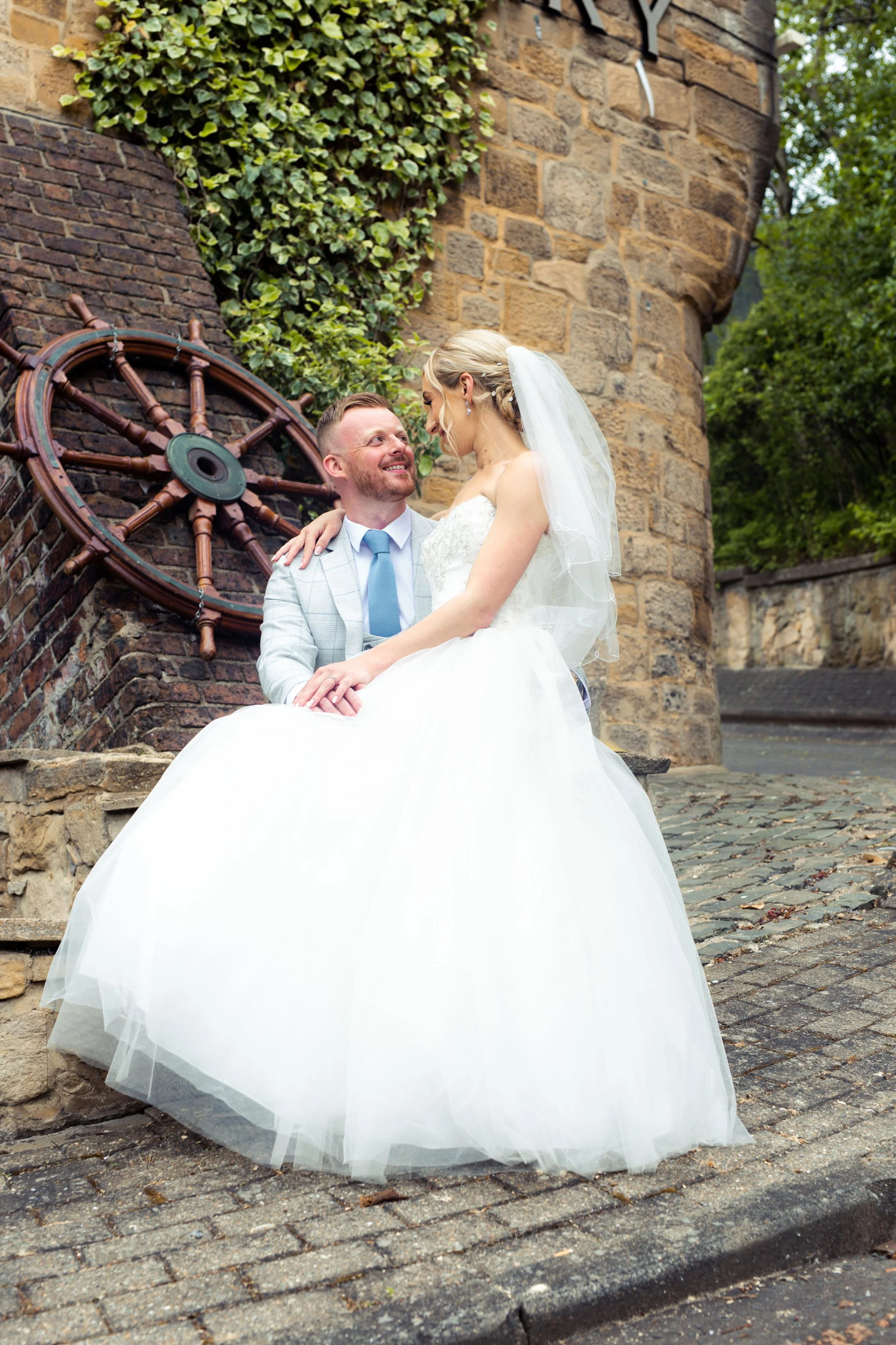 A newlywed couple sitting outdoors on a stone bench in front of a brick wall with a ship's wheel, during daytime, with greenery around.