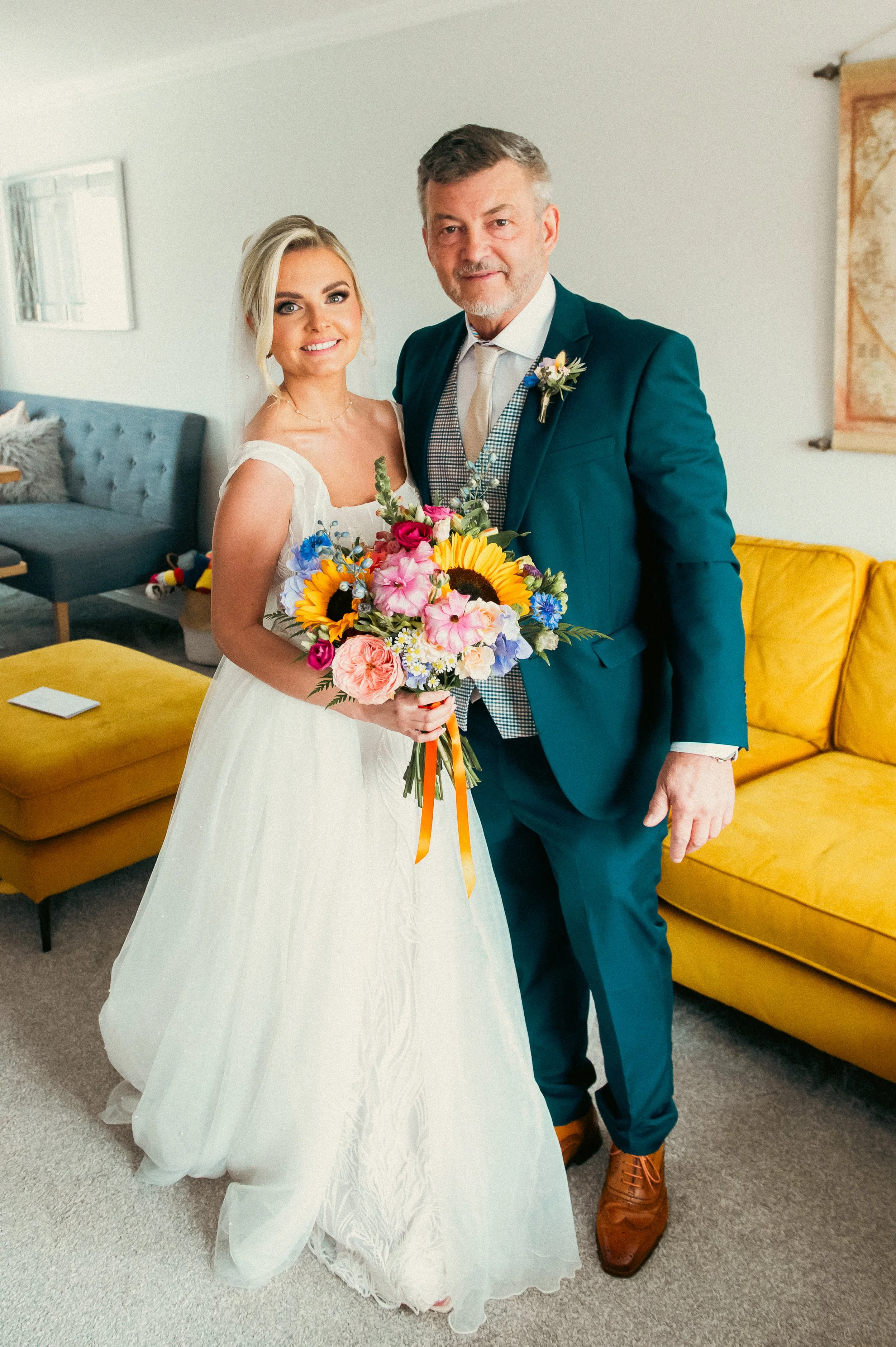 A bride in a white wedding dress holding a vibrant bouquet, standing next to a man in a teal suit inside a living room with yellow couch and gray chair.