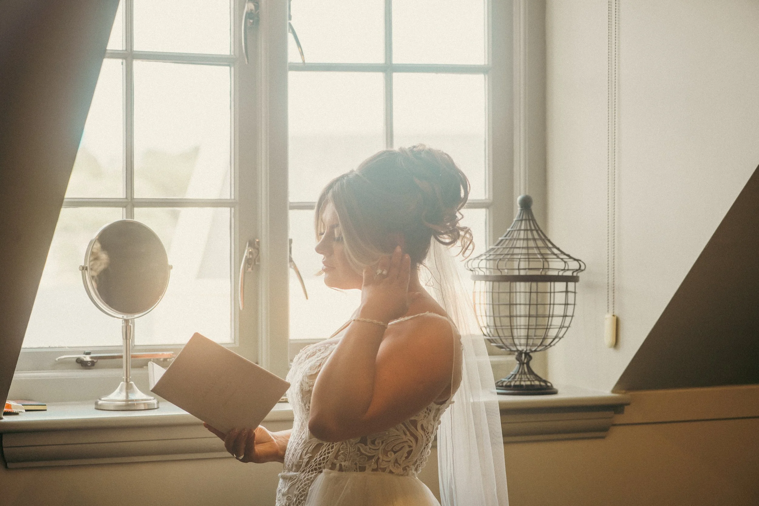 Bridal woman with styled hair, wearing a lace wedding dress, getting ready in front of a window, holding a book and adjusting her earring.