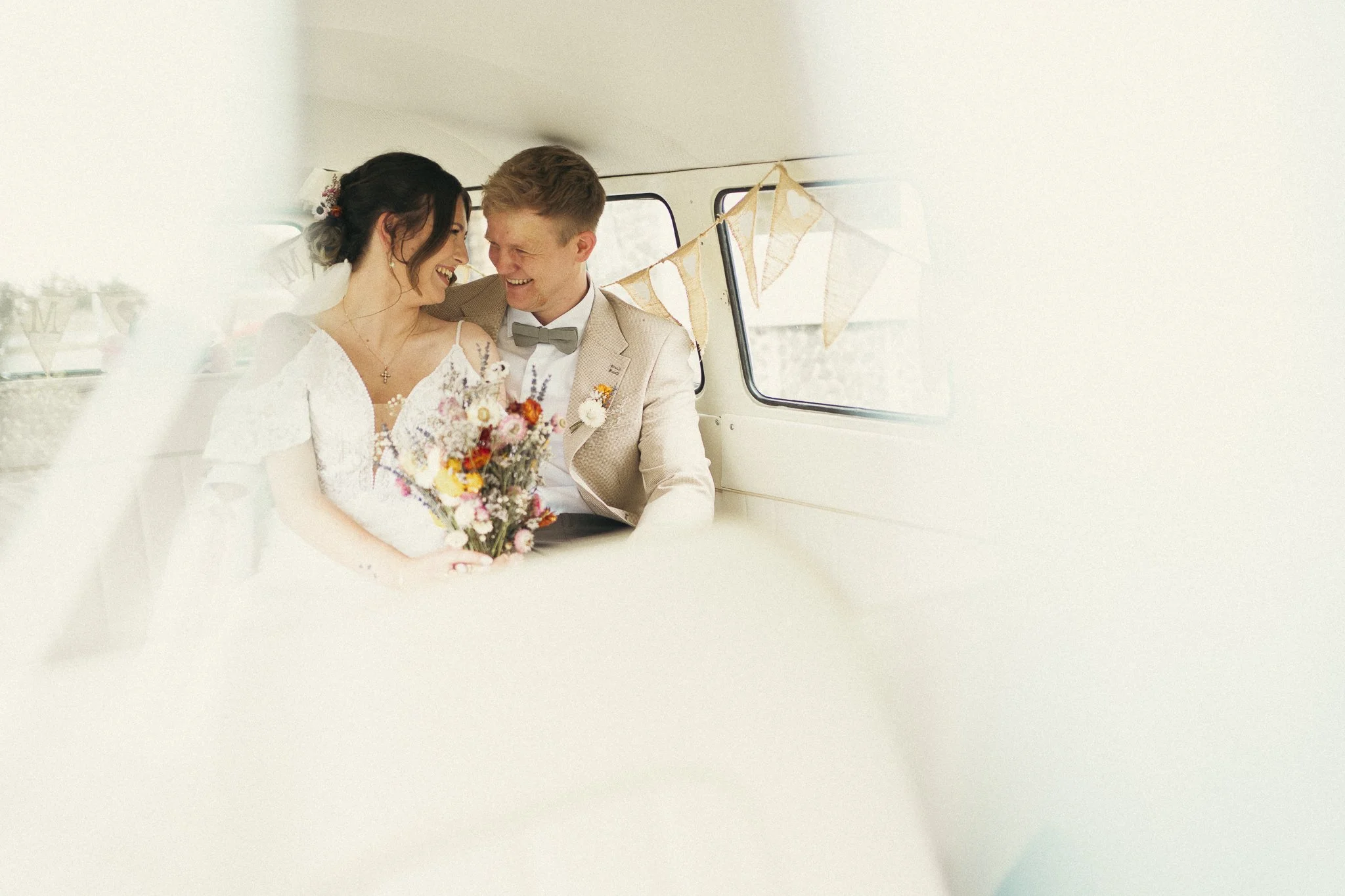 A bride and groom sitting close together inside a decorated vehicle, smiling and looking at each other, with the bride holding a colorful bouquet of flowers.