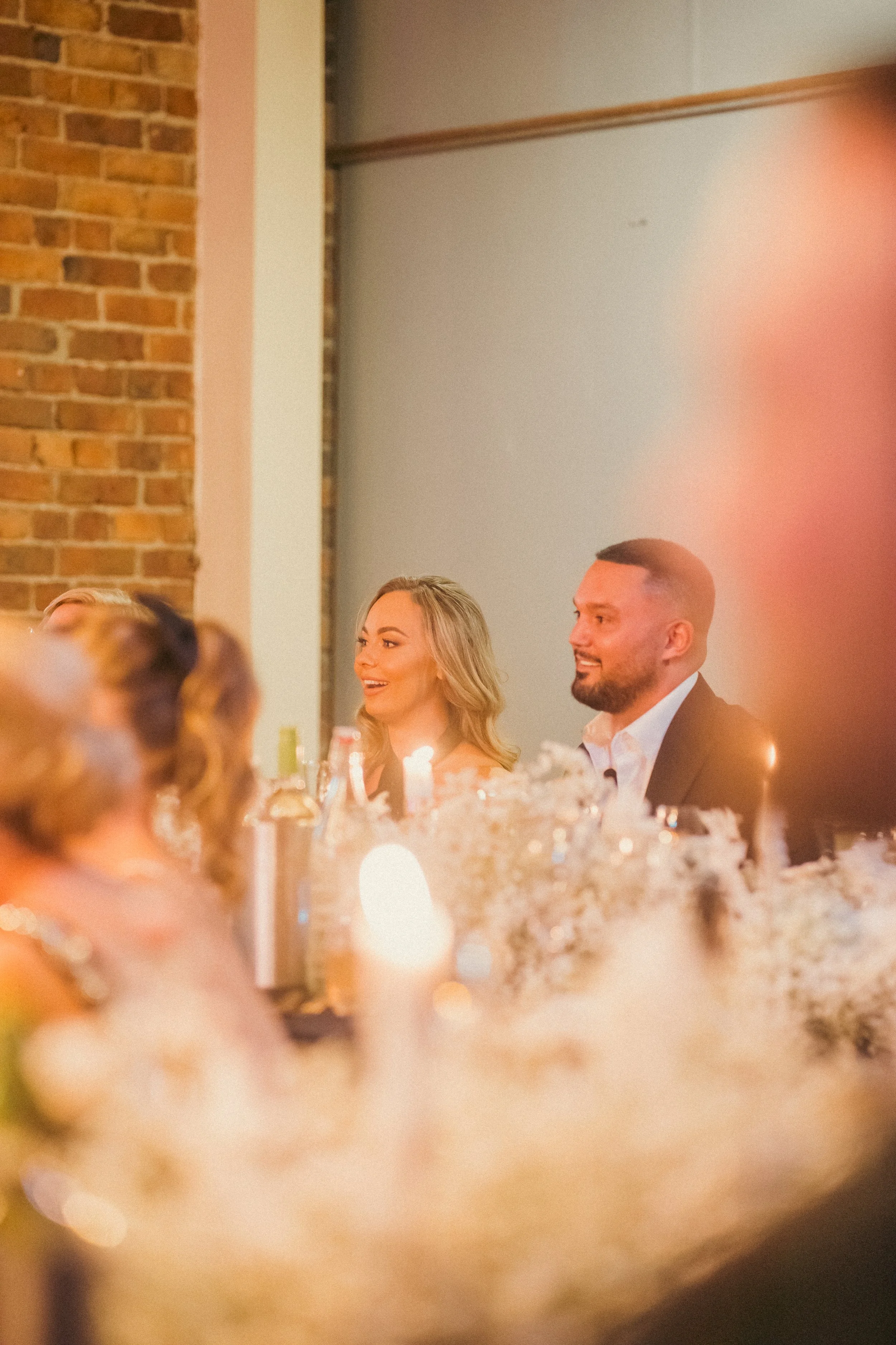 A woman and a man sitting at a dinner table during a celebration, smiling and looking to their left, with candles and floral decorations on the table, and a brick wall and plain wall in the background.