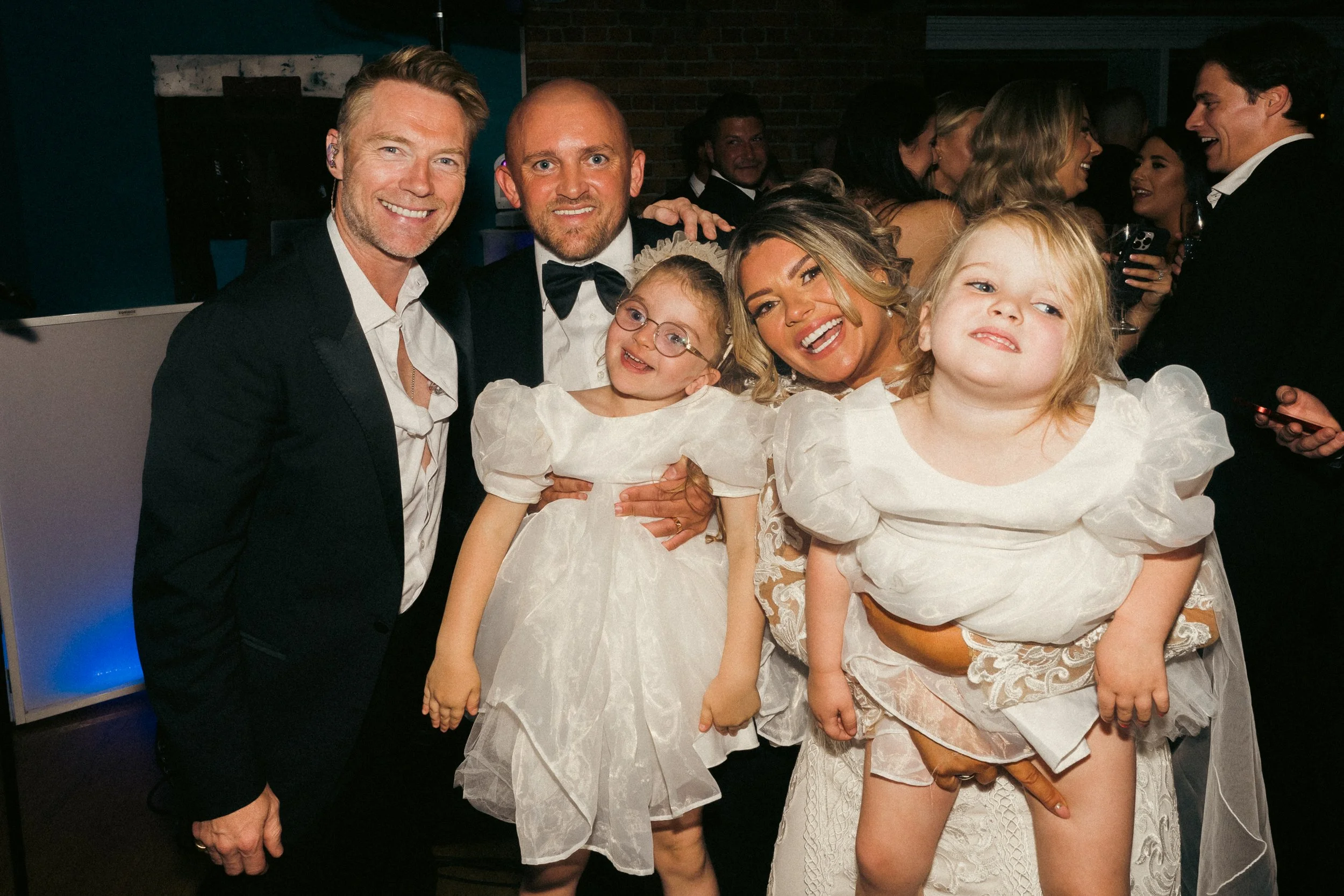 Group of adults and children dressed in formal attire at a celebration or wedding reception, smiling and posing for the photo.