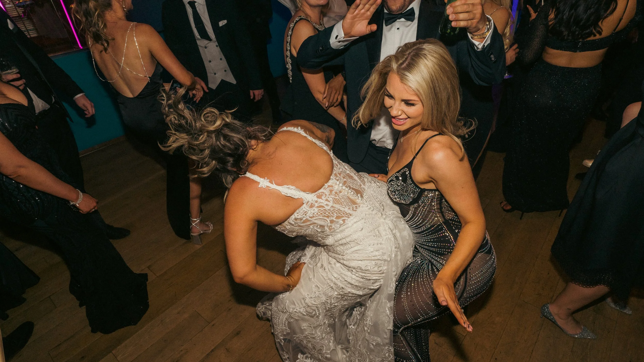 People dancing at a party, with one woman in a white lace dress and another woman in a sparkly, black dress with spaghetti straps dancing closely together, surrounded by other partygoers in fashionable attire.