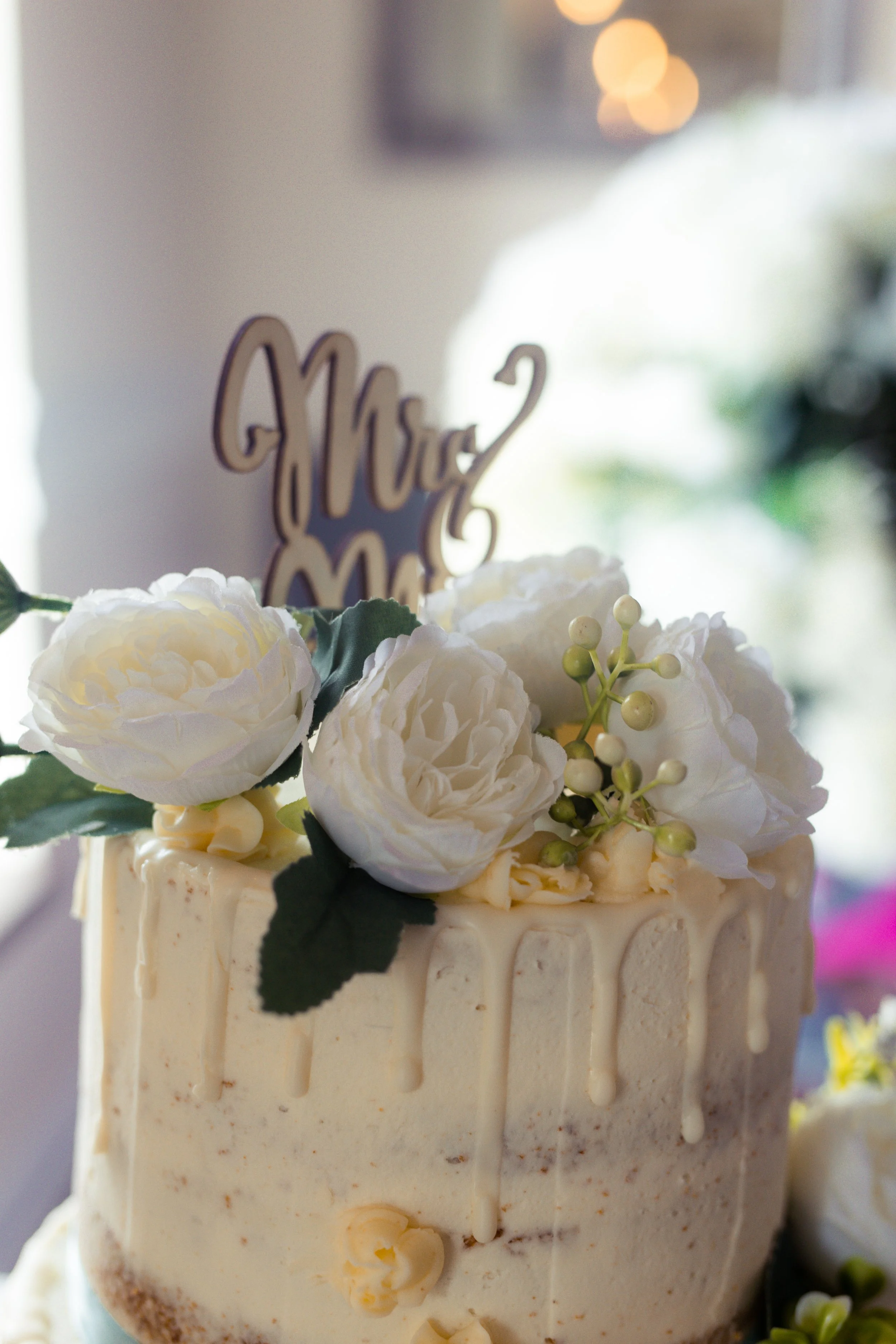 A wedding cake decorated with white flowers and a cake topper that reads 'Mr & Mrs' underneath a blurred background.