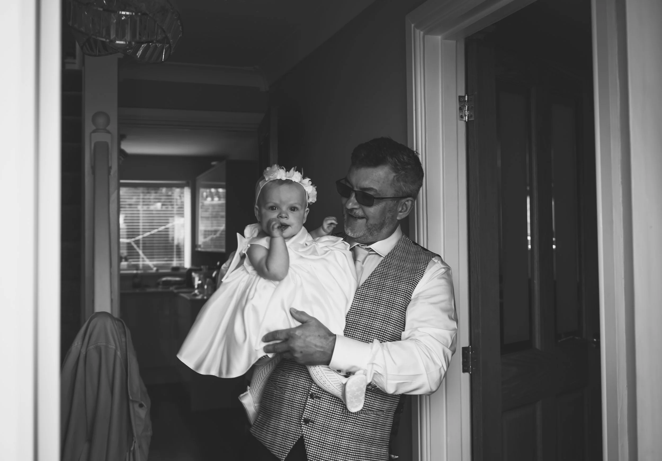 A man wearing sunglasses and a vest holds a young girl in a white dress and floral headband, standing in a home interior near a doorway.