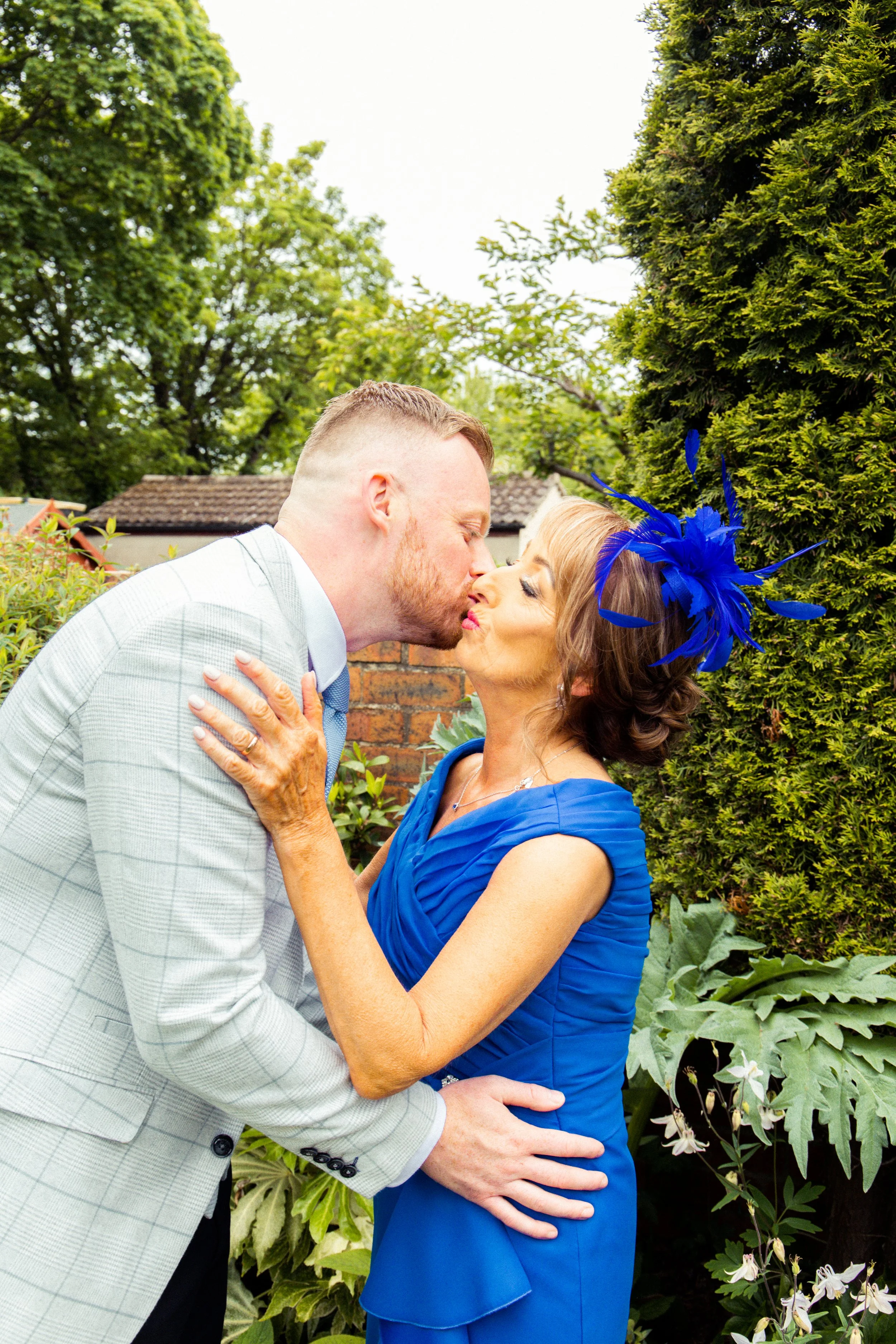 A man and woman sharing a kiss outdoors, with the man in a light gray checked suit and the woman in a vibrant blue dress with a matching blue feathered hat, surrounded by greenery.