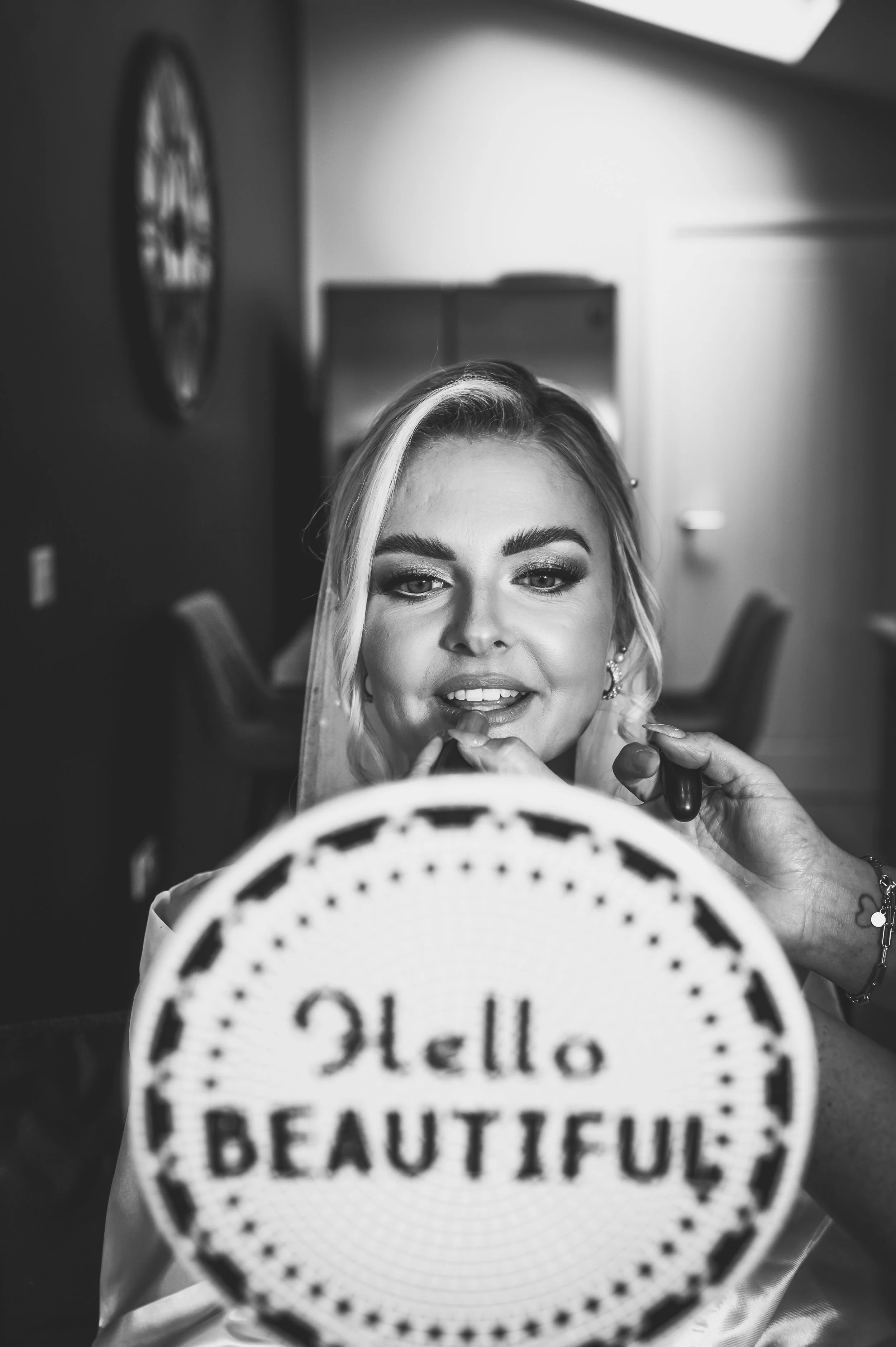 Black and white photo of a woman getting her makeup done, with a sign reading 'Hello BEAUTIFUL' in front of her.