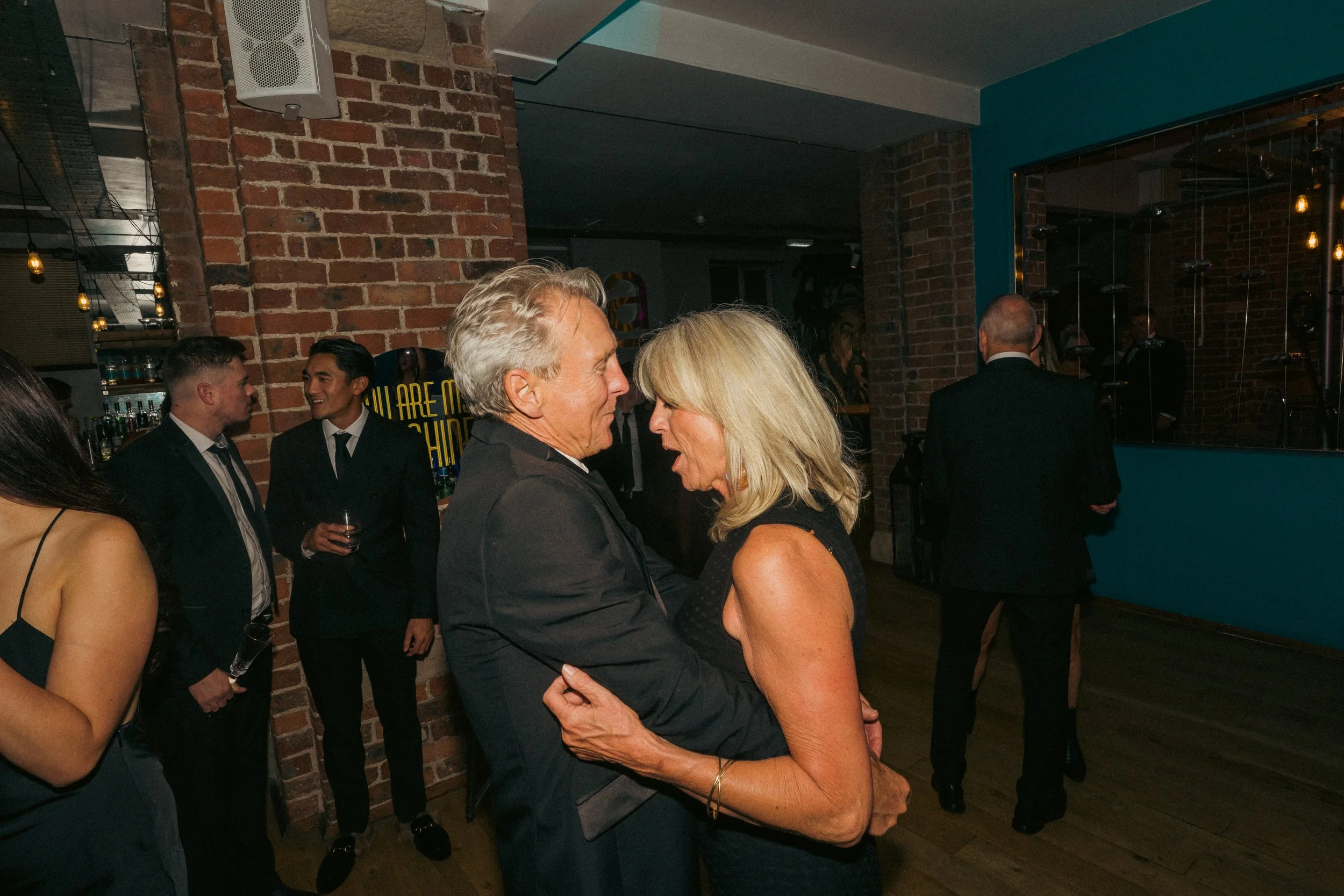 A couple is dancing and talking at an indoor party with brick and teal walls; other guests are socializing in the background.