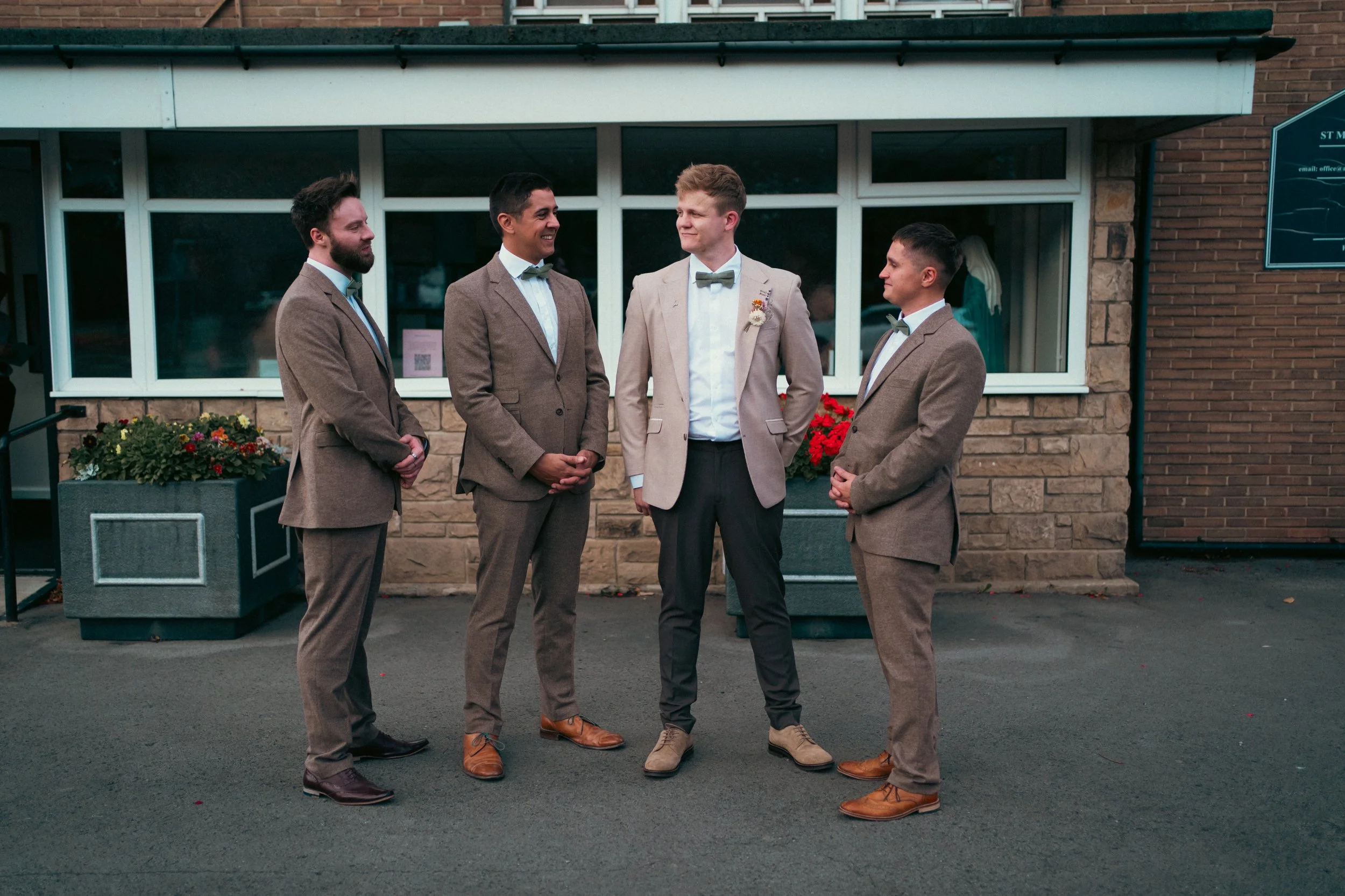 Four men dressed in suits and bow ties standing outside in front of a brick building, engaged in conversation at a social gathering.
