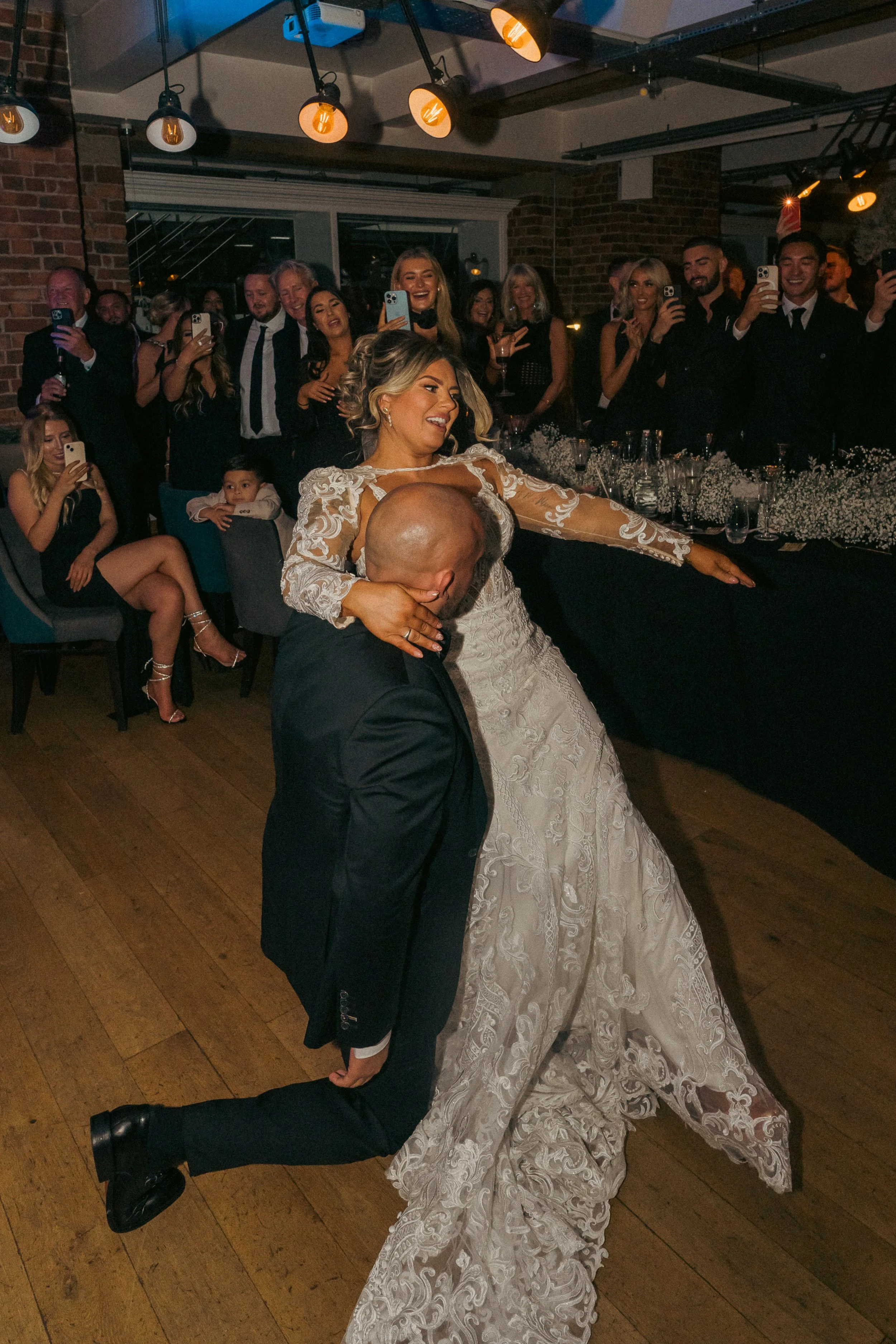 A bride and groom dancing at their wedding reception, with guests watching and taking photos.