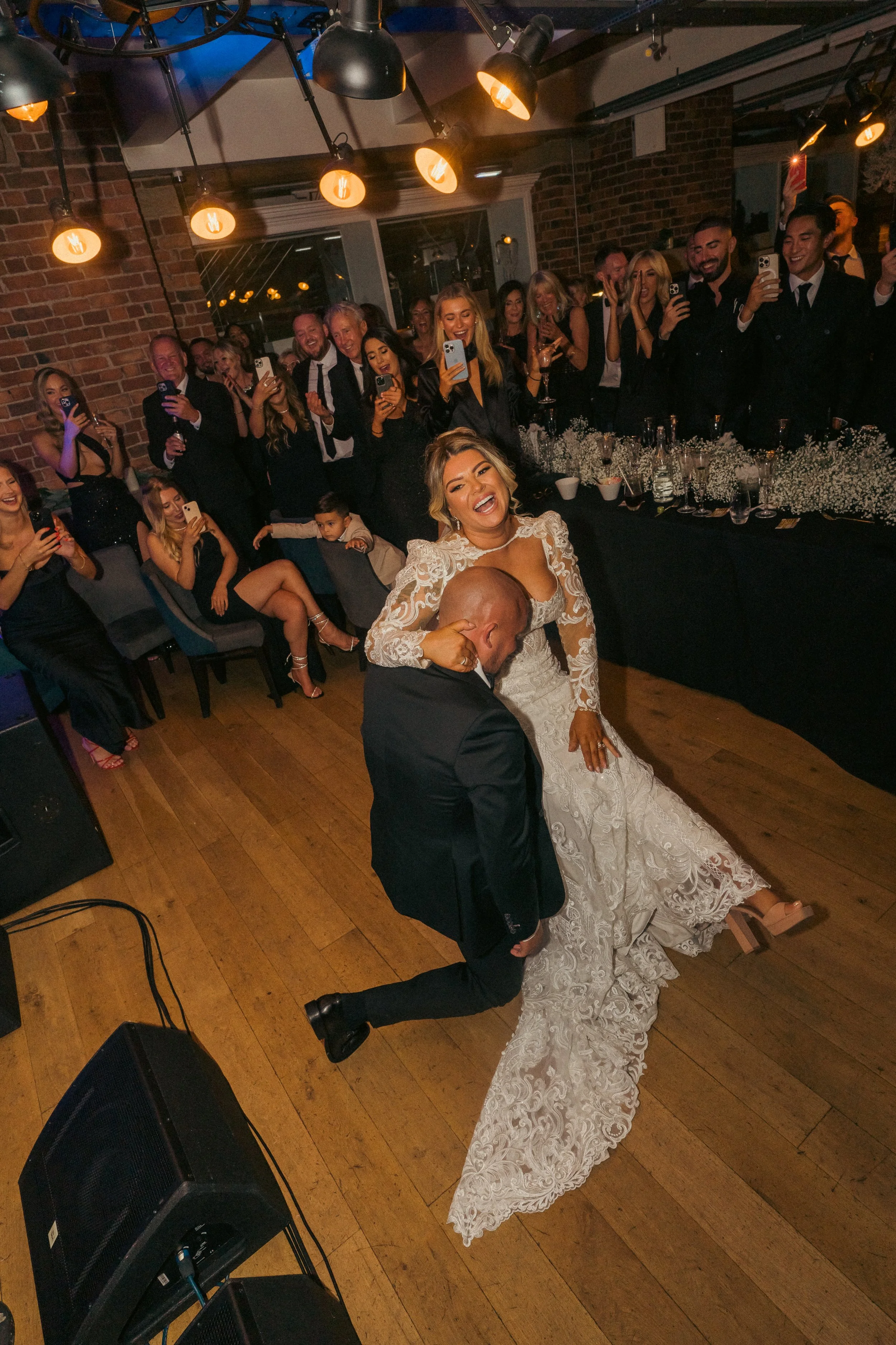 Bride and groom dancing at wedding reception with guests watching and taking photos, in an indoor venue with brick walls and warm lighting.