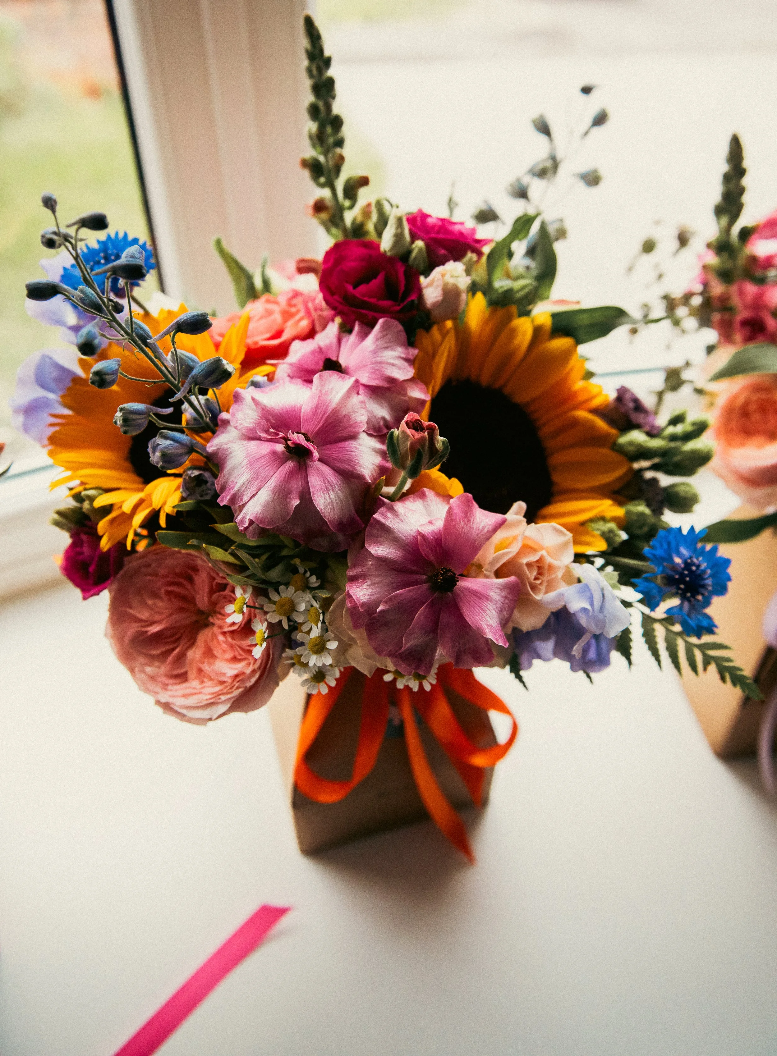 Close-up of a colorful bouquet of flowers, including sunflowers, pink roses, purple flowers, and assorted greenery, in a black vase with an orange ribbon, placed near a window.