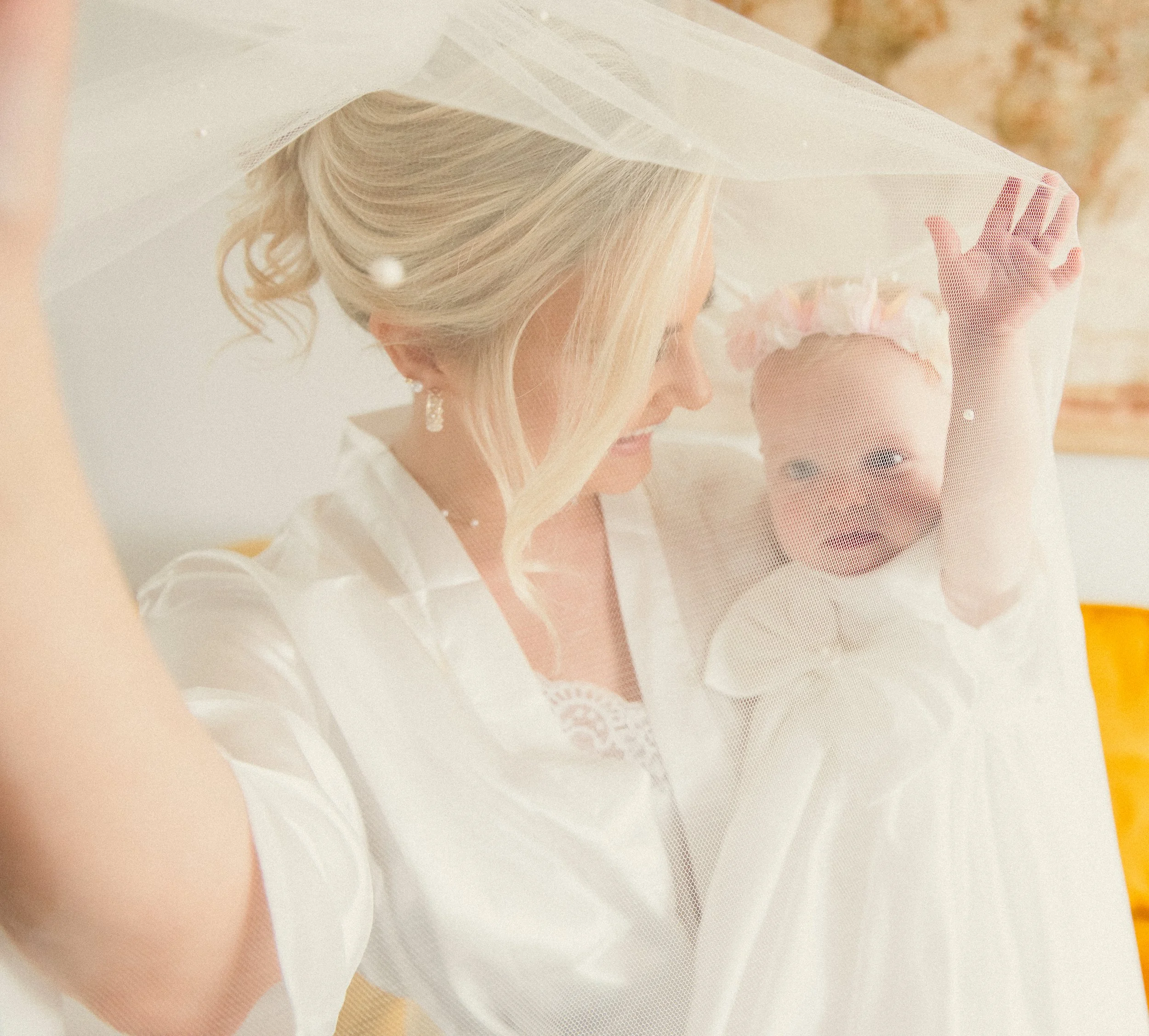 A woman and a baby girl behind a veil, smiling at each other. The woman has blonde hair and is wearing earrings, and the baby girl is wearing a white dress and a floral headband.