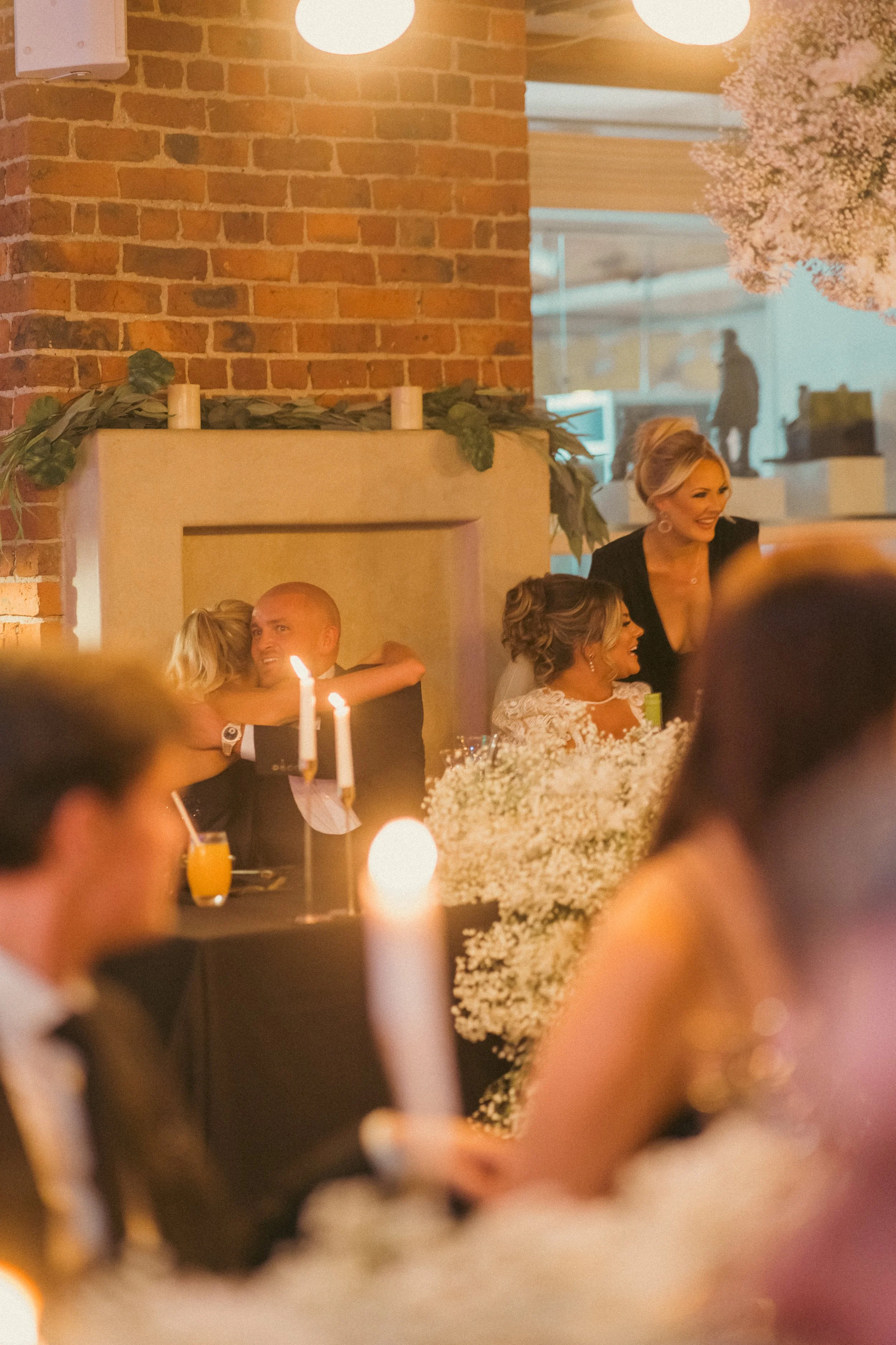 A group of people celebrating at a wedding reception, with candles on the table, some hugging, smiling, and chatting, with a brick wall and floral decorations in the background.