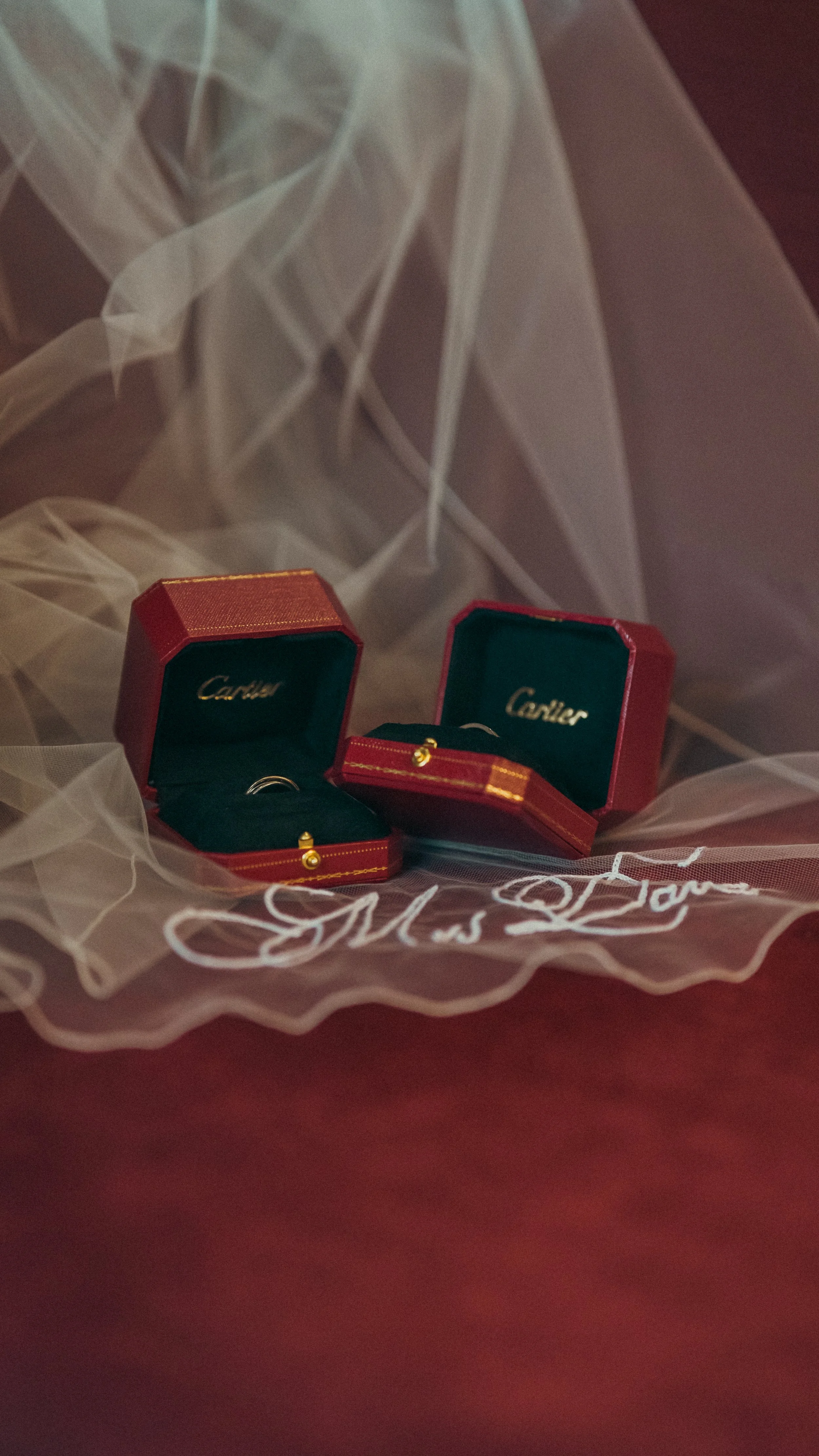 Two Cartier ring boxes on a tulle cloth with white embroidery, one open revealing a ring, and a gold wedding band next to the boxes.