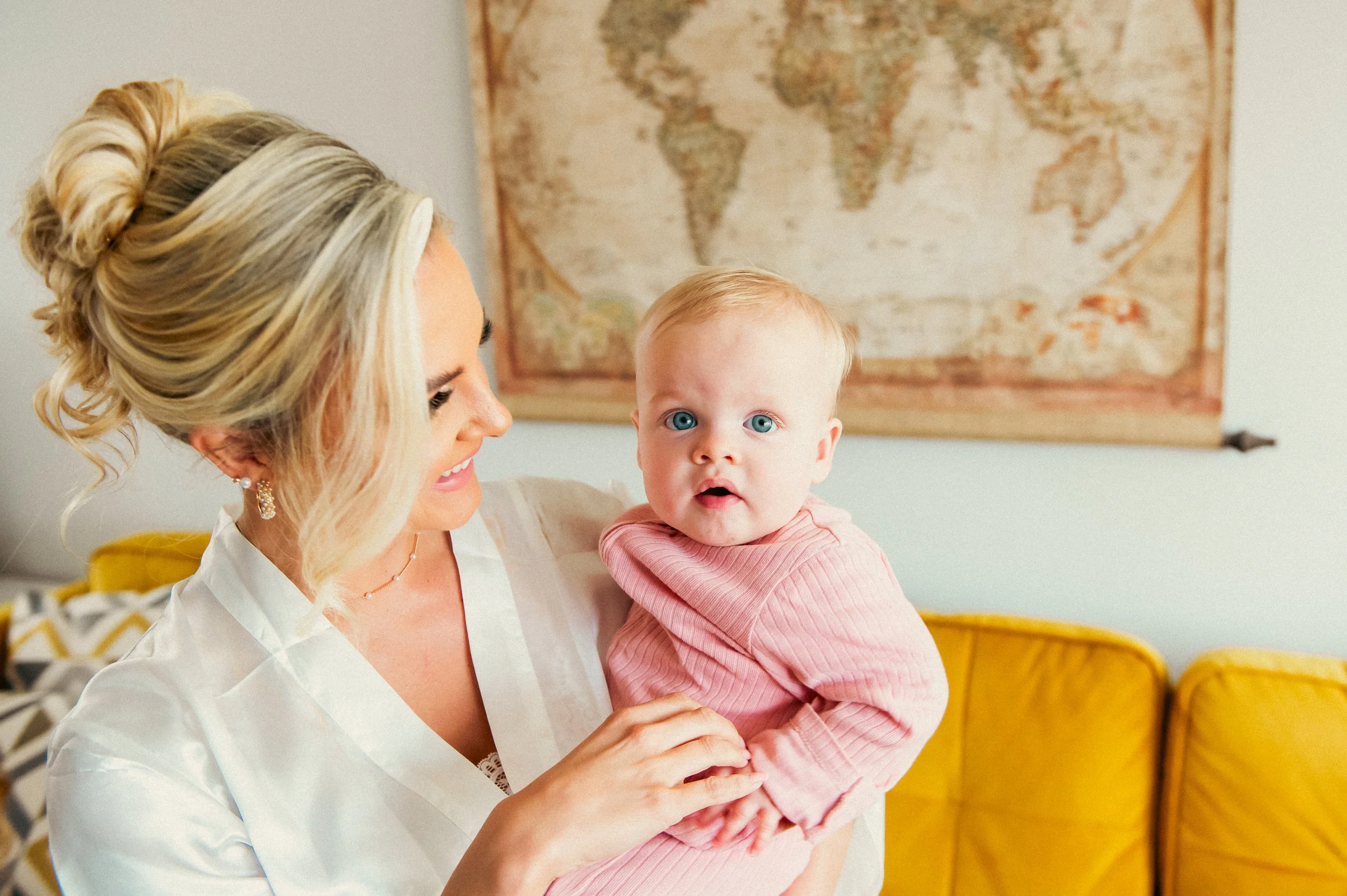 A woman with blonde hair styled in an updo holding a baby with blonde hair and blue eyes, against a backdrop of a world map on the wall.