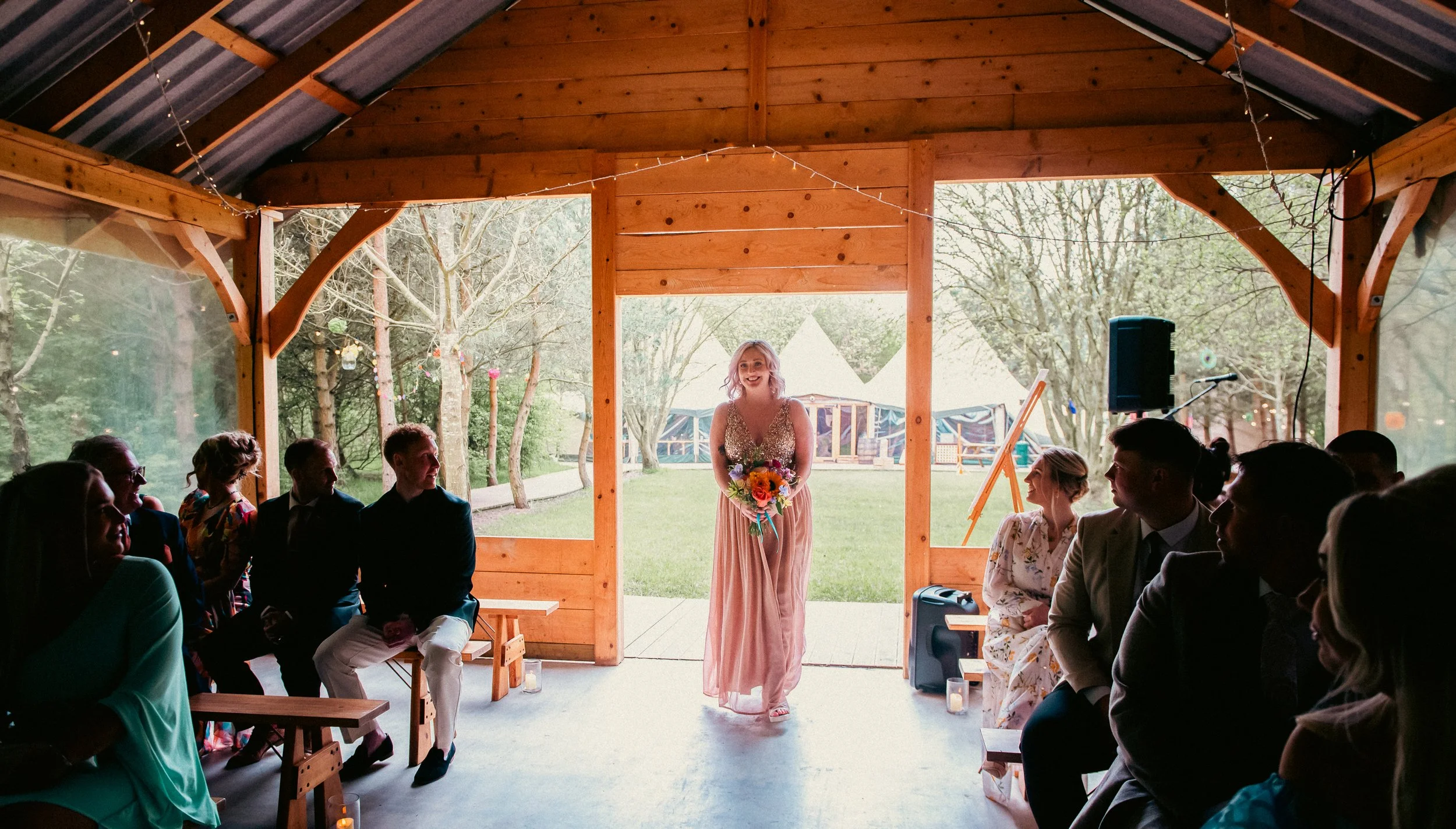 A woman in a sleeveless, sparkly top and peach-colored skirt stands with a bouquet of flowers in a wooden pavilion during a wedding ceremony, with guests seated on either side and trees outside.