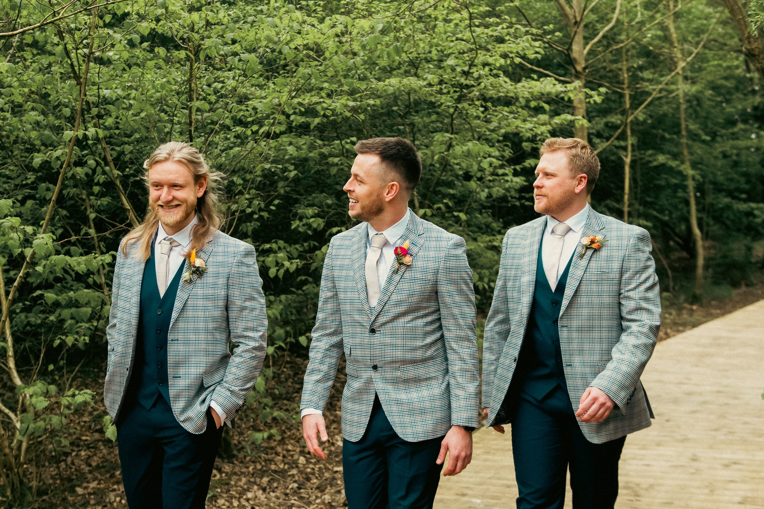 Three men in matching plaid blazers, blue vests, and white shirts walking outdoors on a trail, with lush green trees in the background, smiling and engaging in conversation.