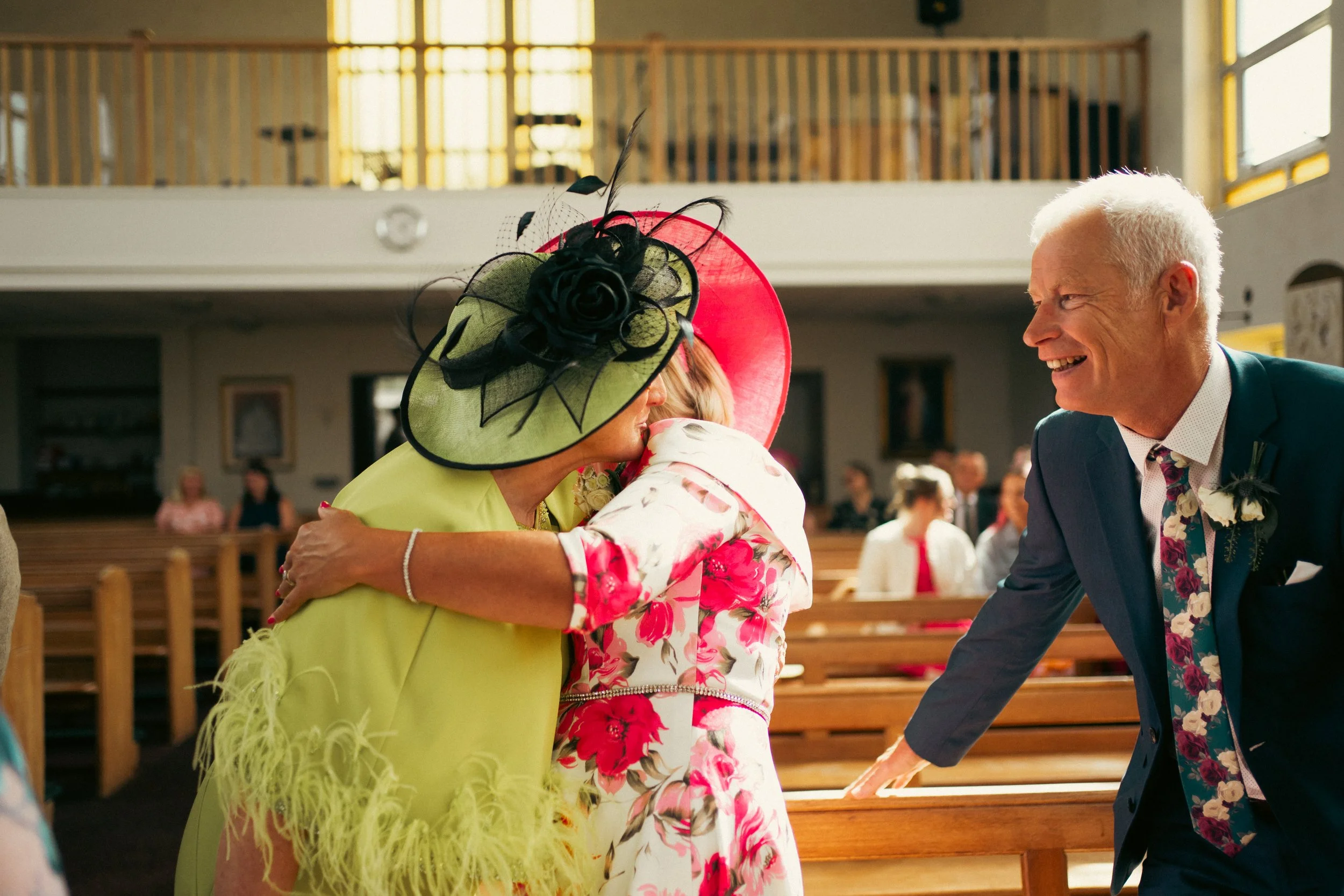 A wedding scene inside a church with three people in focus: a woman in a yellow dress with a large green and black hat, hugging an older woman in a floral dress with a large red hat, and a man in a blue suit with a floral tie smiling and leaning forw
