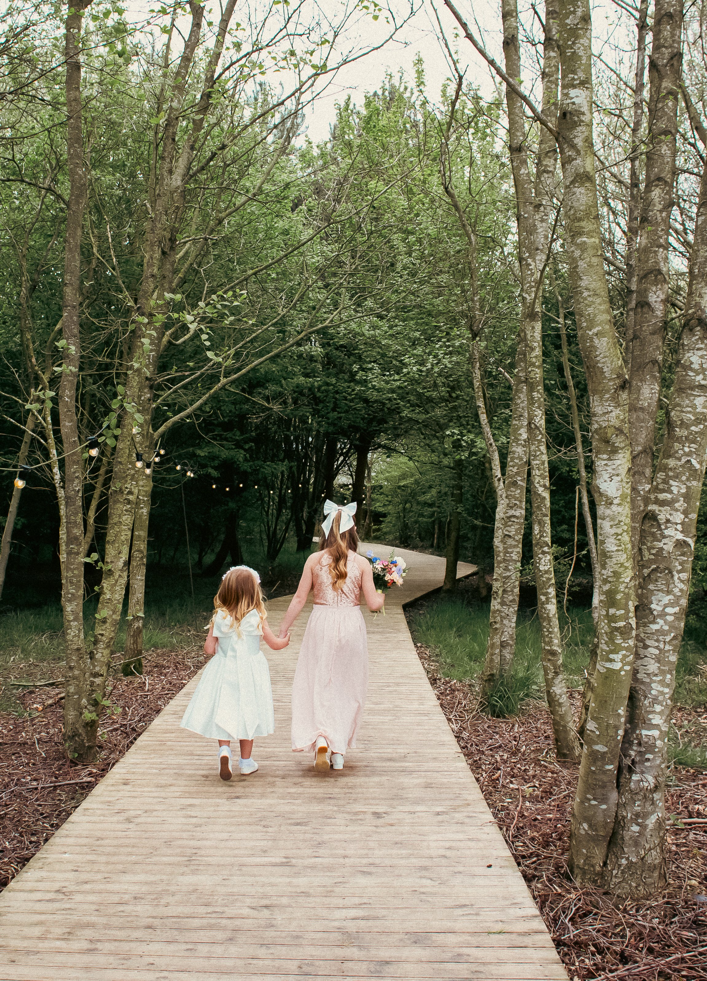 Two girls walking hand in hand on a wooden trail through a forest, one holding a bouquet of flowers.