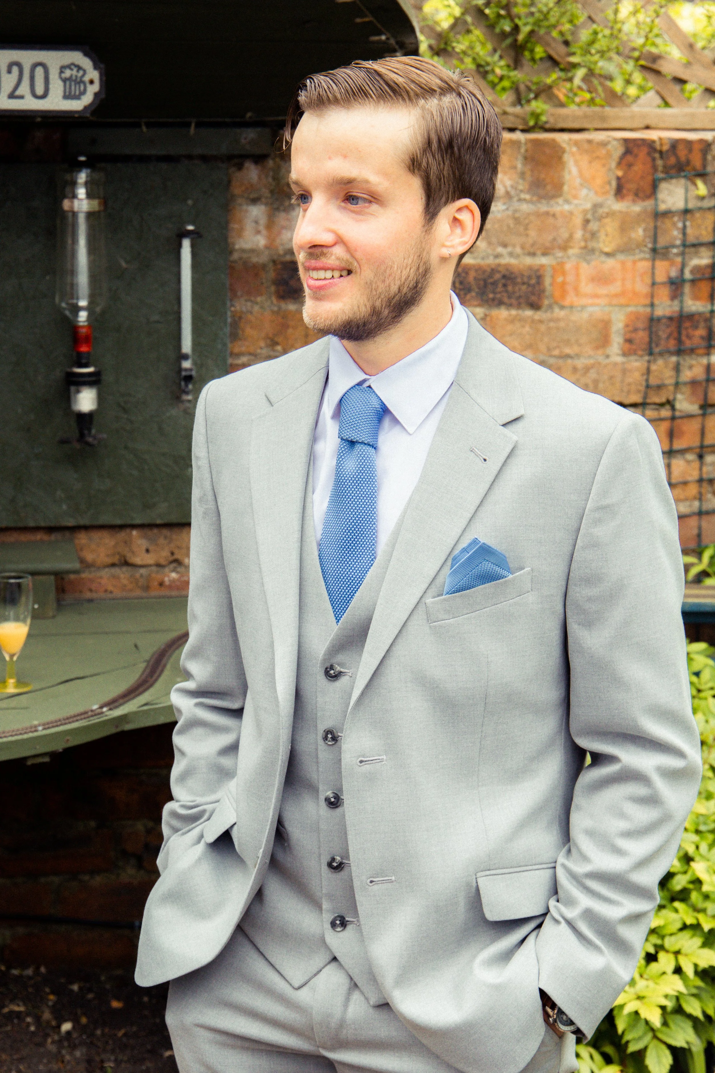 A young man wearing a light gray suit with a blue tie and matching pocket square, standing outdoors with a brick wall and greenery in the background.