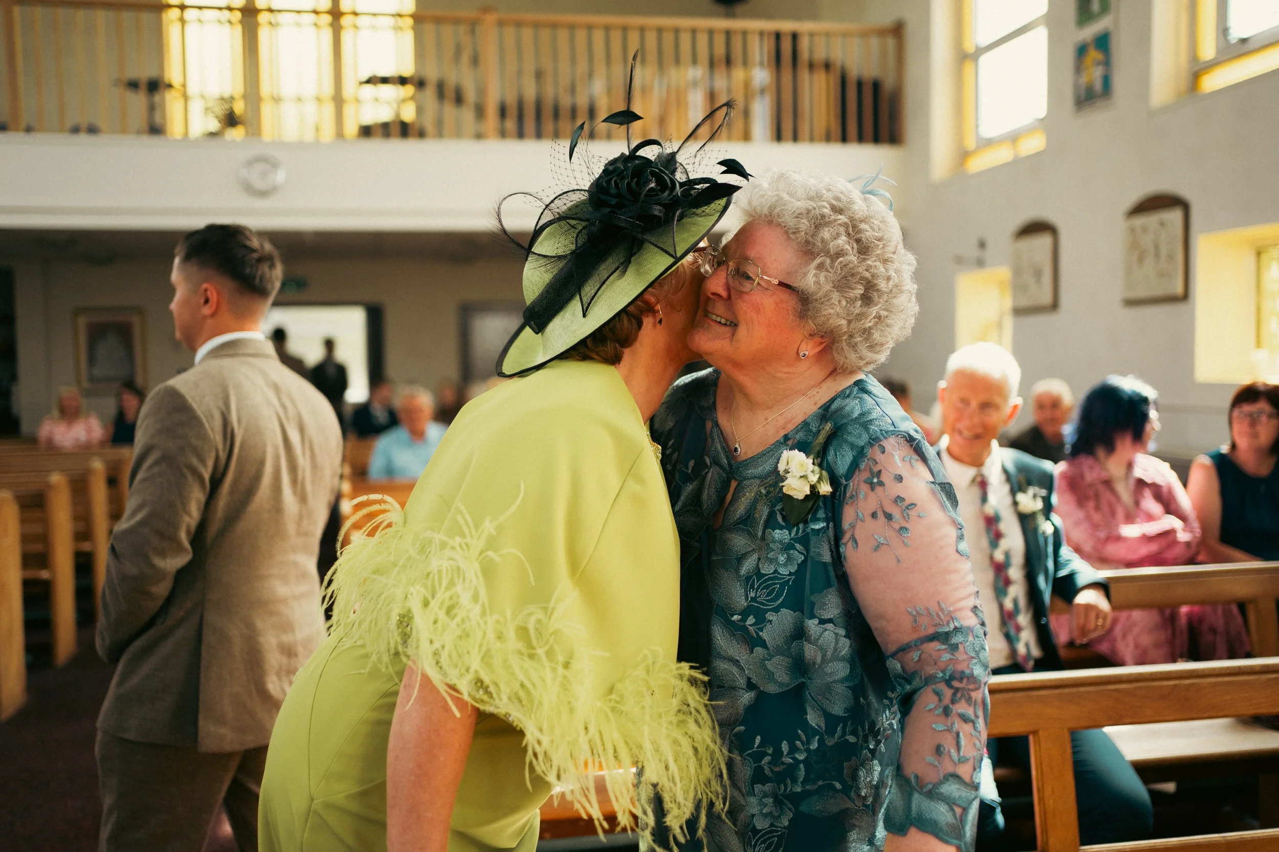 Two women, one in a bright yellow dress and large hat, and the other in a blue lace dress, greeting each other warmly with kisses on the cheek inside a church during a wedding ceremony. Other guests are seated in the background.