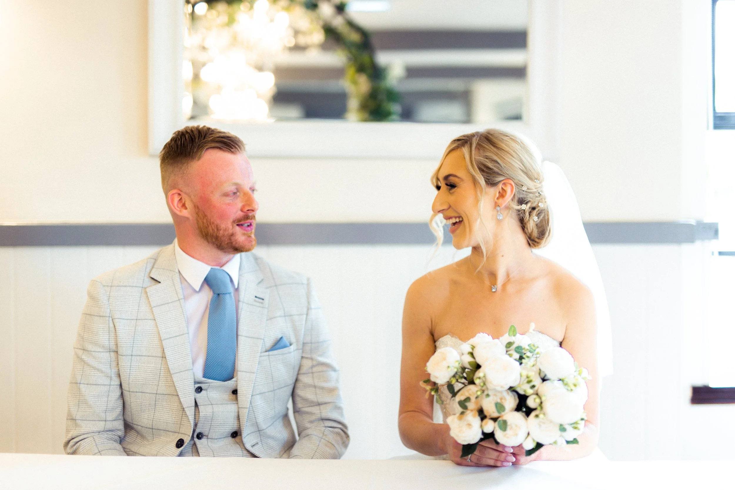 A bride and groom sitting at a table during their wedding ceremony, smiling at each other. The bride is holding a bouquet of white flowers and wearing a strapless wedding gown, while the groom is dressed in a light-colored checked suit with a blue ti